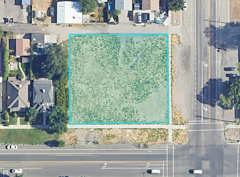 Aerial view of a vacant lot with sparse grass, surrounded by residential houses on the left and a busy street on the right.