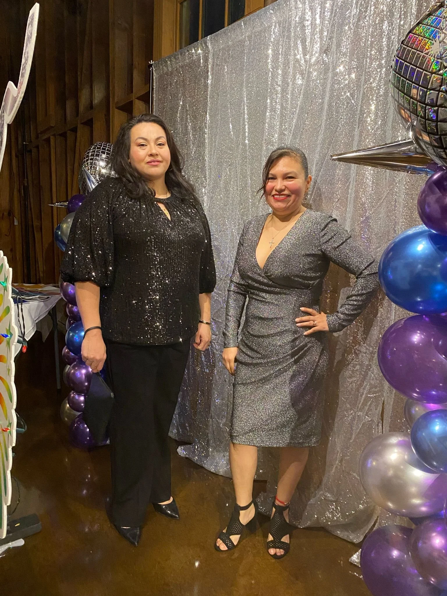 Two women dressed in sparkly and elegant outfits stand in front of a silver, glittery backdrop with colorful balloons on the sides, at a festive celebration or party.