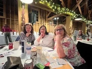 Three women sitting at a decorated table during a gathering or celebration in a barn-like setting with string lights and rustic decor.