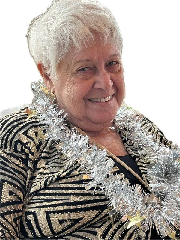 An elderly woman with short white hair, smiling cheerfully, wearing a black and gold patterned jacket and silver tinsel garland around her neck.