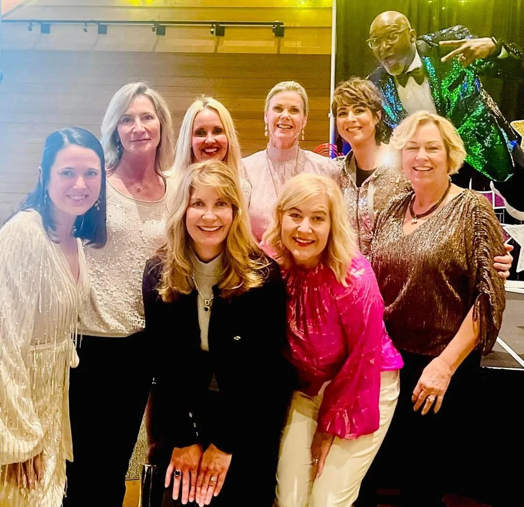 Group of eight women standing together at a celebration event, with one man in a colorful sparkling blazer in the background. The women are dressed in festive attire, smiling at the camera.