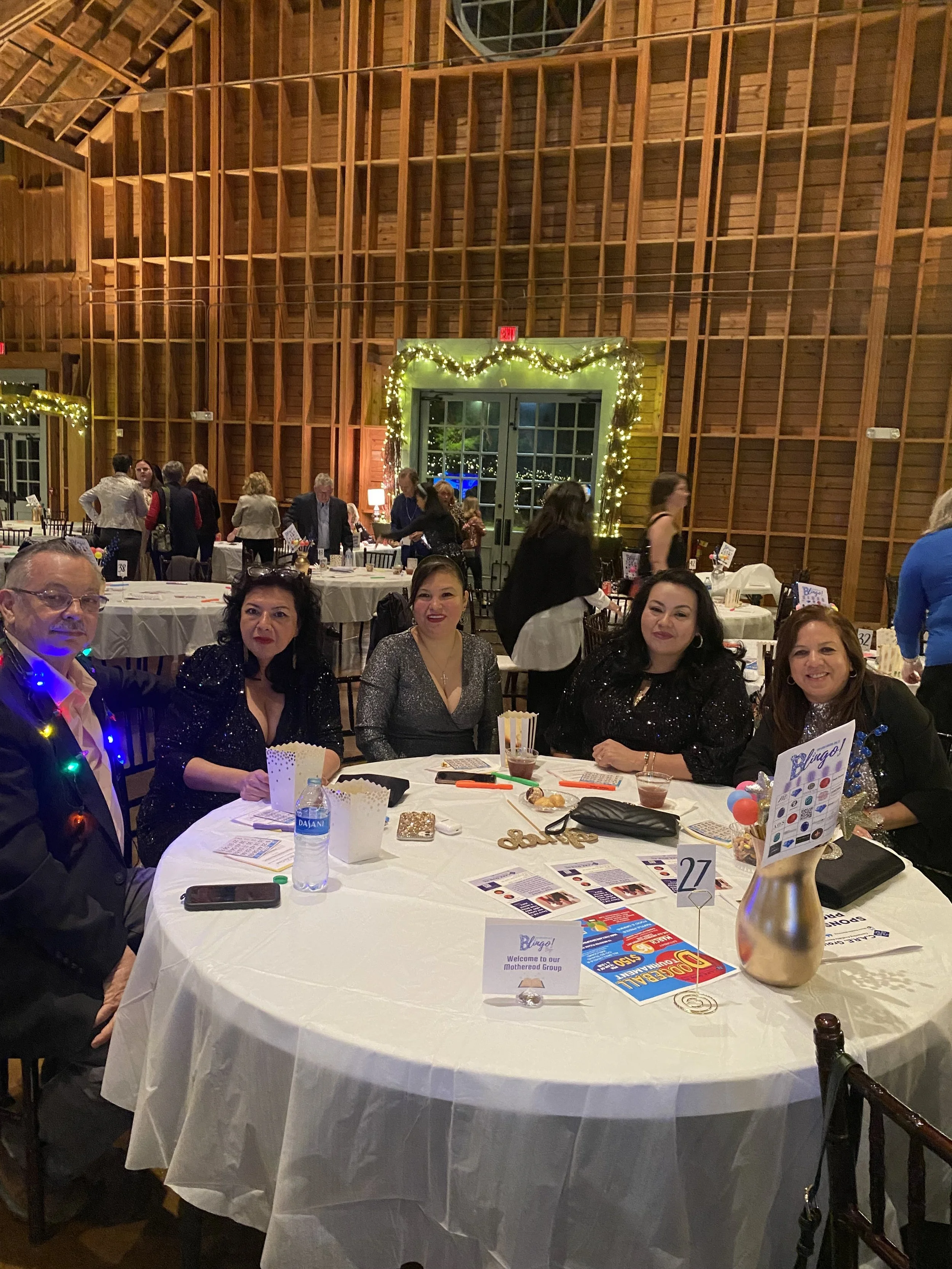 Group of five people sitting at a round table decorated for a celebration, with holiday lights and a wooden wall in the background.