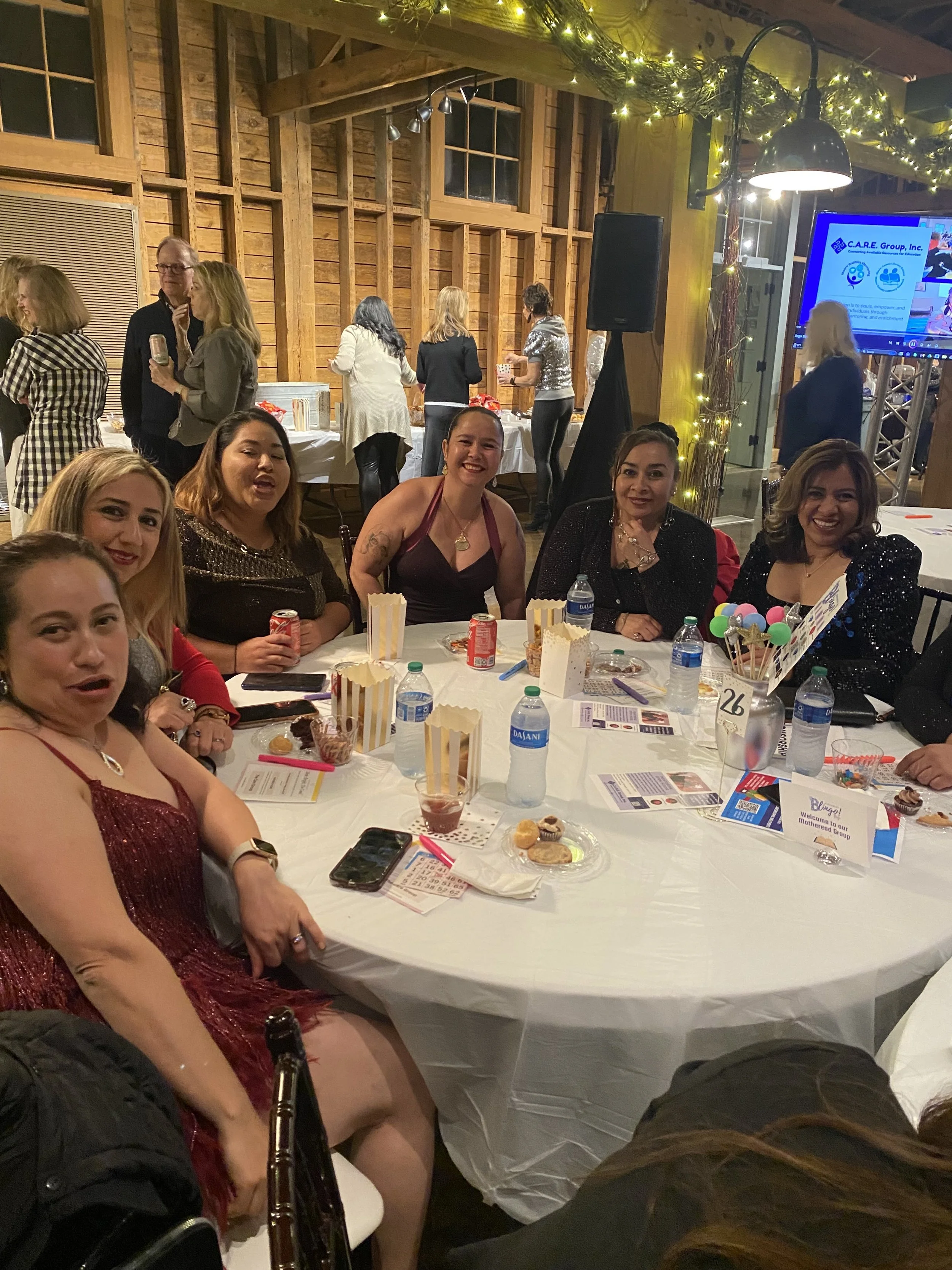 Group of women sitting around a table at a party with snacks, drinks, and decorations in a wooden venue with a banner and a screen in the background.