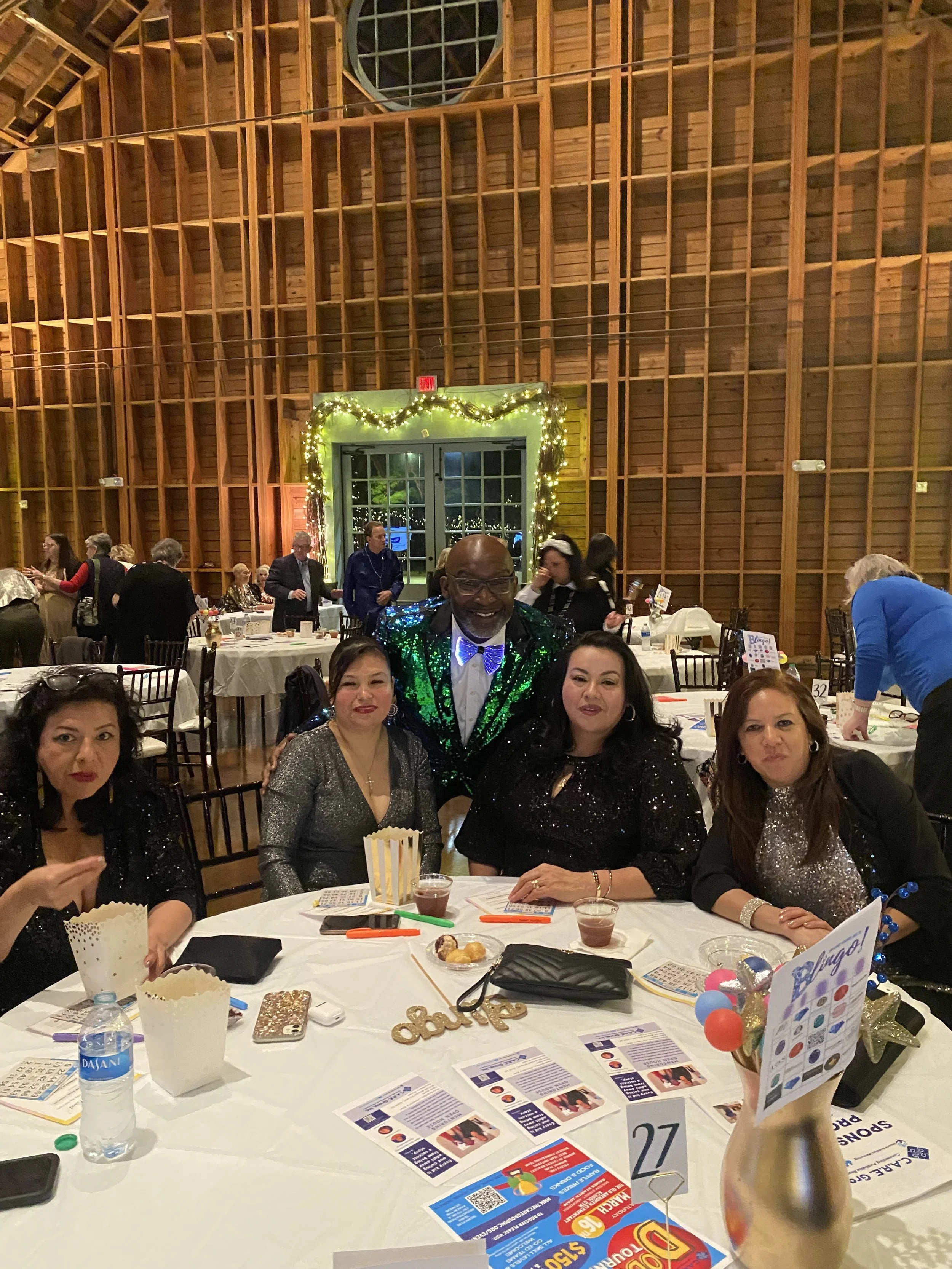 Group of five people sitting and standing around a table at a festive gathering in a wooden hall decorated with fairy lights.