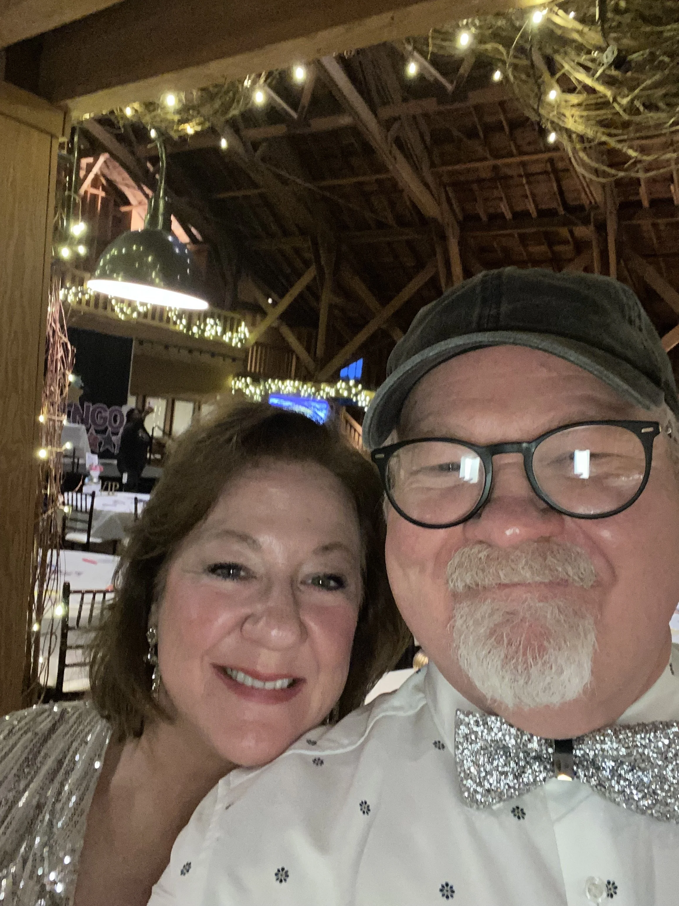 A smiling woman and man taking a selfie together at an indoor event, decorated with string lights and a stage in the background.