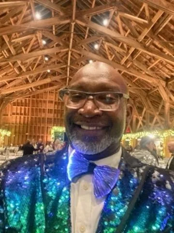 A man wearing glasses and a shiny, colorful jacket with a bow tie, smiling inside a wooden structure with a high, arched ceiling at an event or gathering.