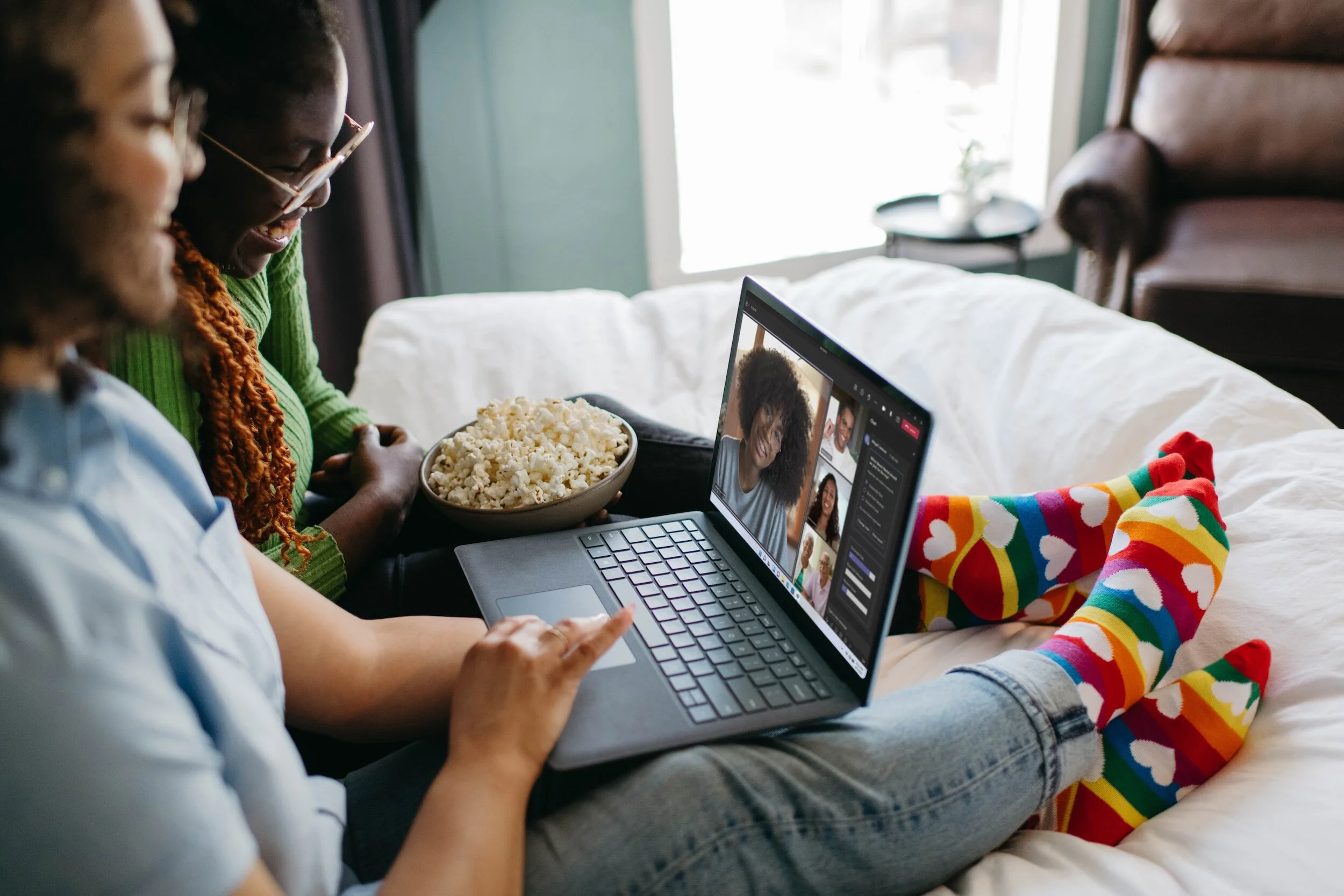 Two women with colorful socks sitting on a white couch, watching a video call on a laptop, with one woman holding a bowl of popcorn.