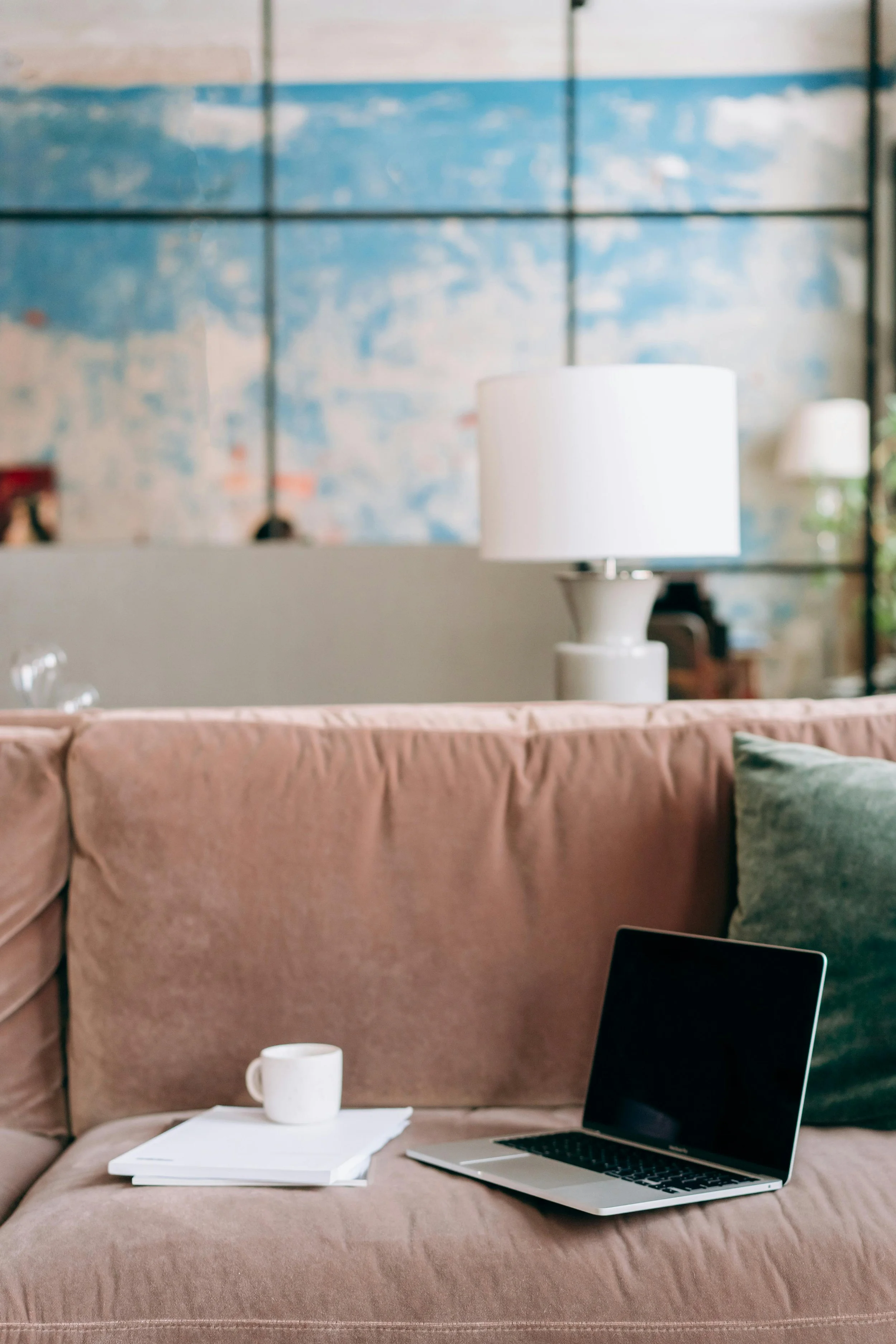 A pink sofa with a green pillow, a laptop, a white mug, and some paper on it, in a cozy living room with an abstract blue and beige wall in the background and a white table lamp.