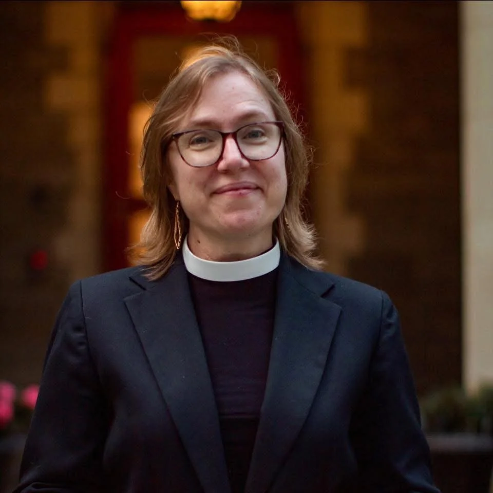 Portrait of a woman with short blonde hair, glasses, and a clerical collar, smiling at the camera against a plain light background.