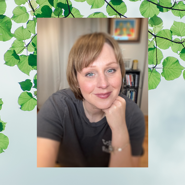 A woman with short blonde hair and blue eyes smiling, resting her chin on her hand, in a gray t-shirt inside a room with a bookshelf and framed artwork in the background.