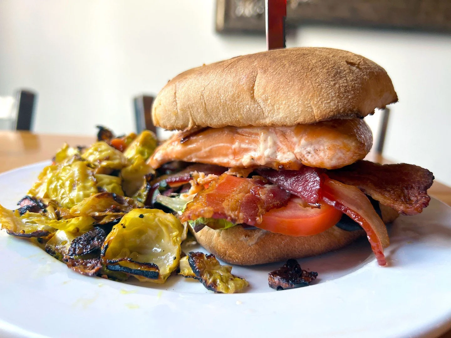 SALMON B.L.T.: grilled salmon, bacon, mixed greens, tomato, fried capers, and deep-fried garlic mayo on a ciabatta bun.Pictured here with a side of ROASTED BRUSSELS SPROUTS. Full menuLINK IN BIO.