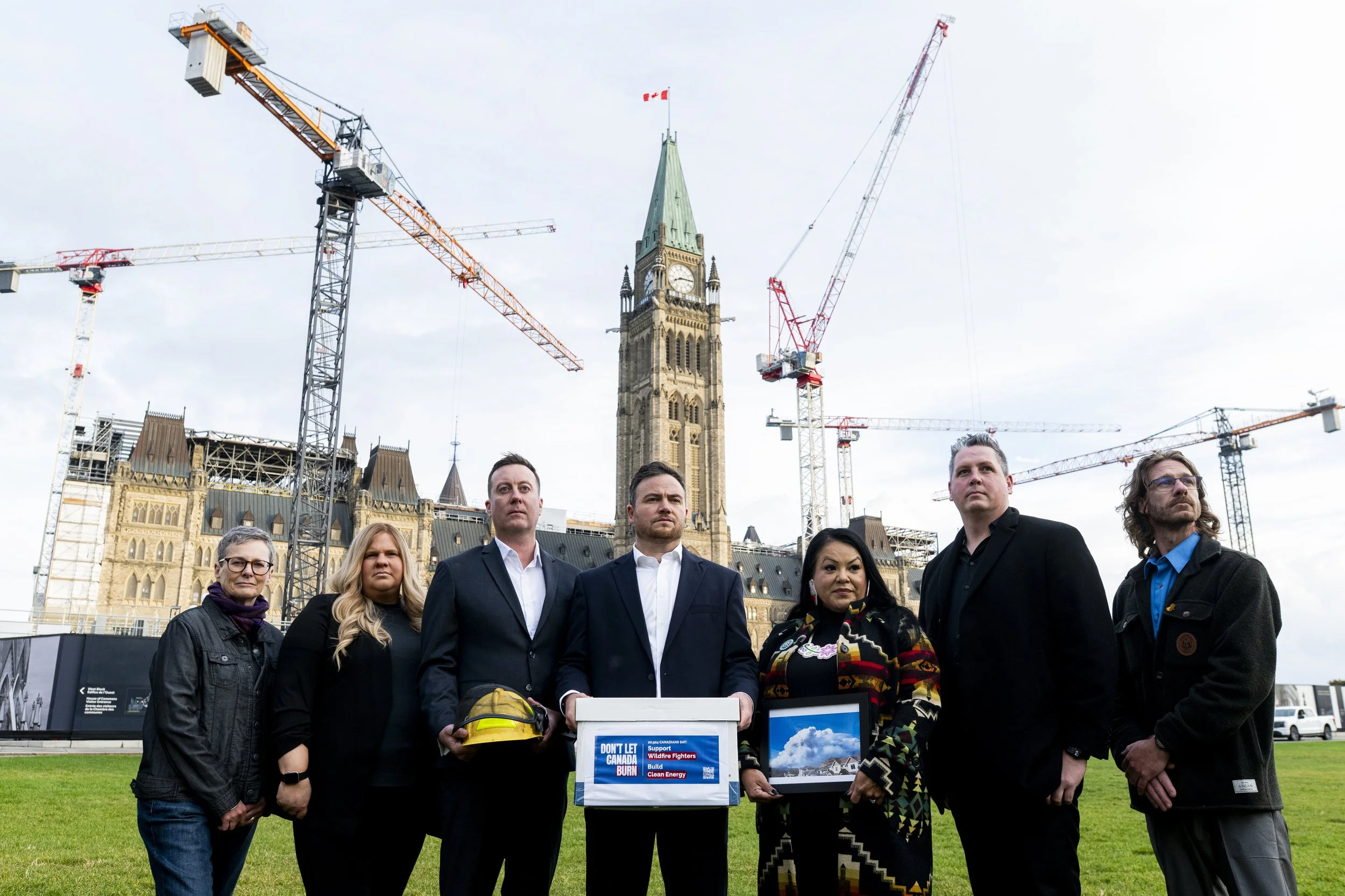 A group of seven people standing on a grassy area in front of the Canadian Parliament buildings in Ottawa, holding signs and pictures, with construction cranes and the Parliament's clock tower in the background.