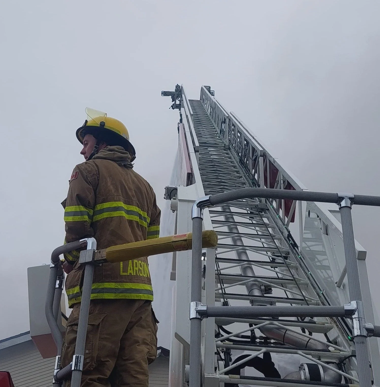 Firefighter standing next to a ladder truck, wearing a yellow helmet and protective gear, with a smoky sky in the background.