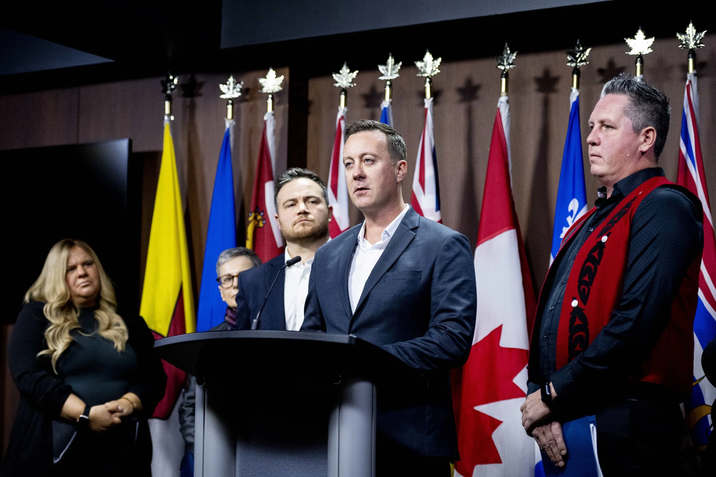 Man in a blue suit speaking at a podium with multiple flags in the background, including a red and white Canadian flag, during a press conference or formal event.