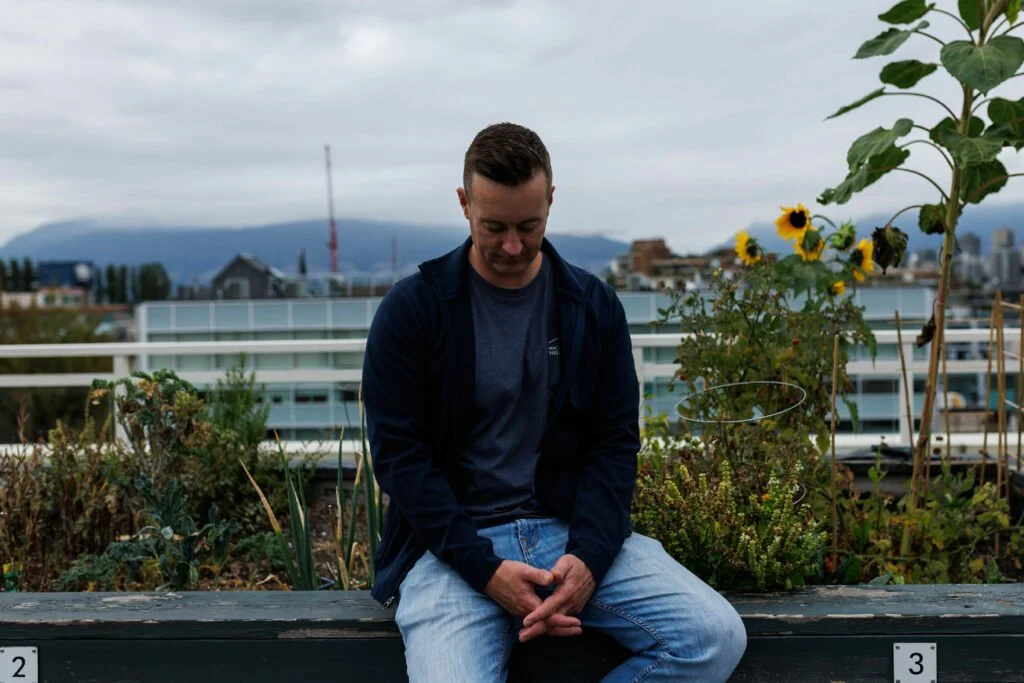 A man sitting on a wooden bench on a rooftop garden with flowers and plants, with city buildings and mountains in the background under cloudy skies.