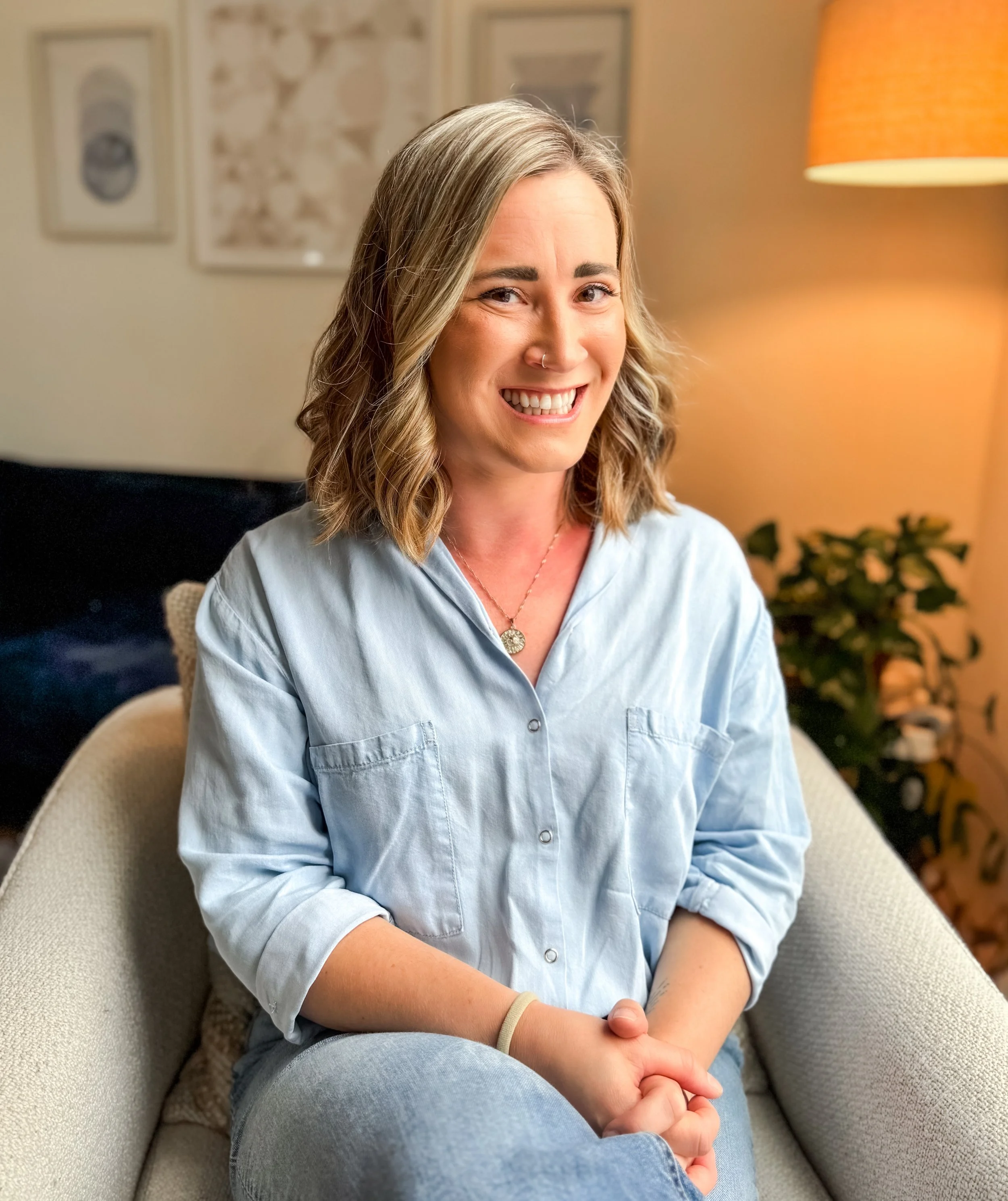 Woman in a white blazer and orange top standing outdoors, wearing blue jeans with a scenic background of trees and mountains.