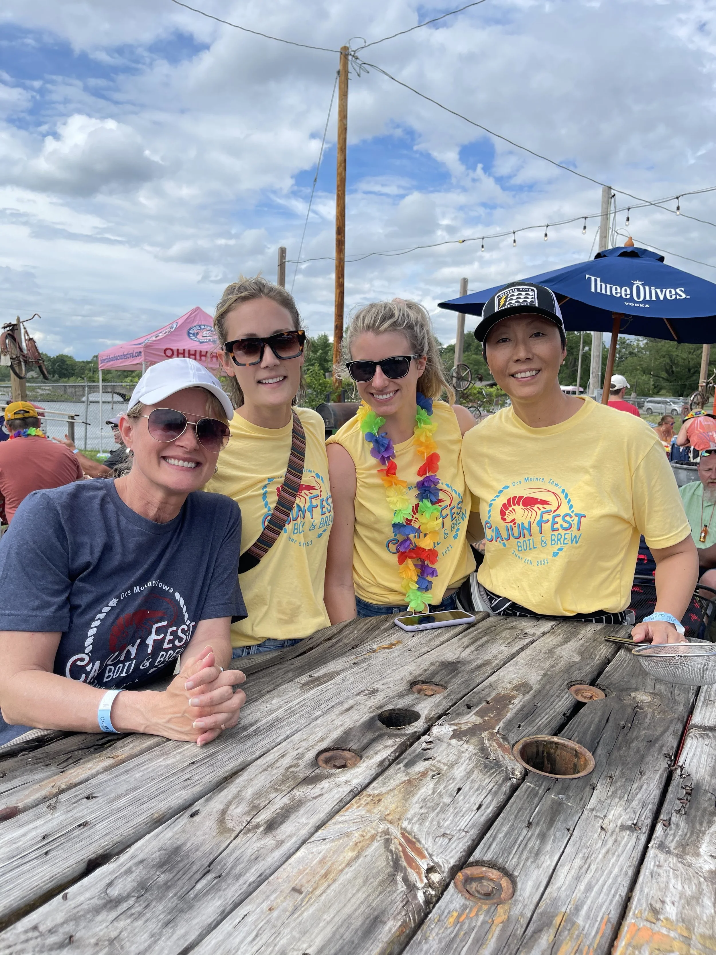 Four people smiling at a rustic outdoor table during a festival. Three of them wear yellow "Cajun Fest Boil & Brew" t-shirts, one with a rainbow lei. The fourth person wears a white cap and a gray t-shirt. String lights and a blue "Three Olives" umbr