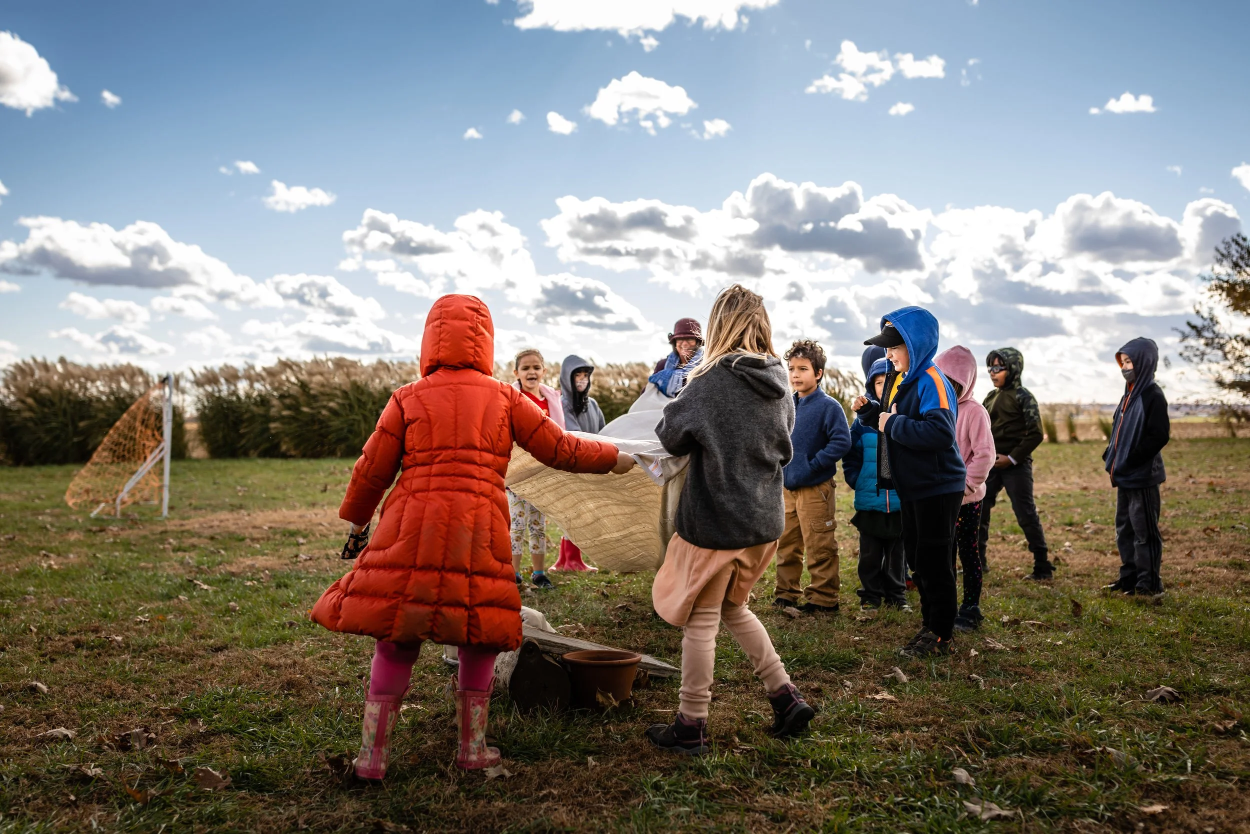 Students play outside with a canvas for a game at the Prairie Moon Waldorf School in Kansas. Wearing red jacket and grey sweater on a soccer field