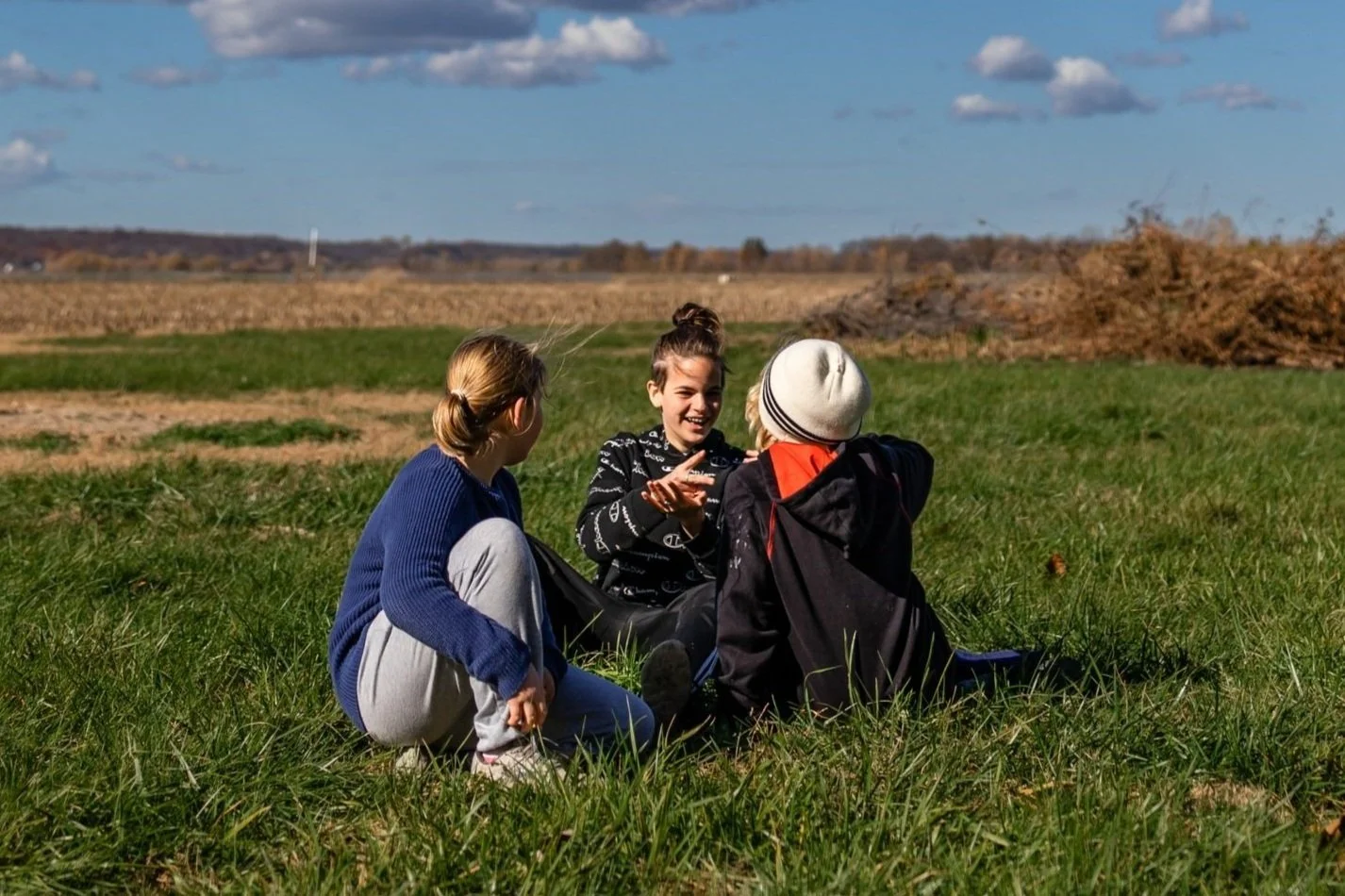 Three 7th grade students sit in a green field outdoors in kansas at the Prairie Waldorf School. They are engaged in conversation and the one visible face is joyful and smiling.