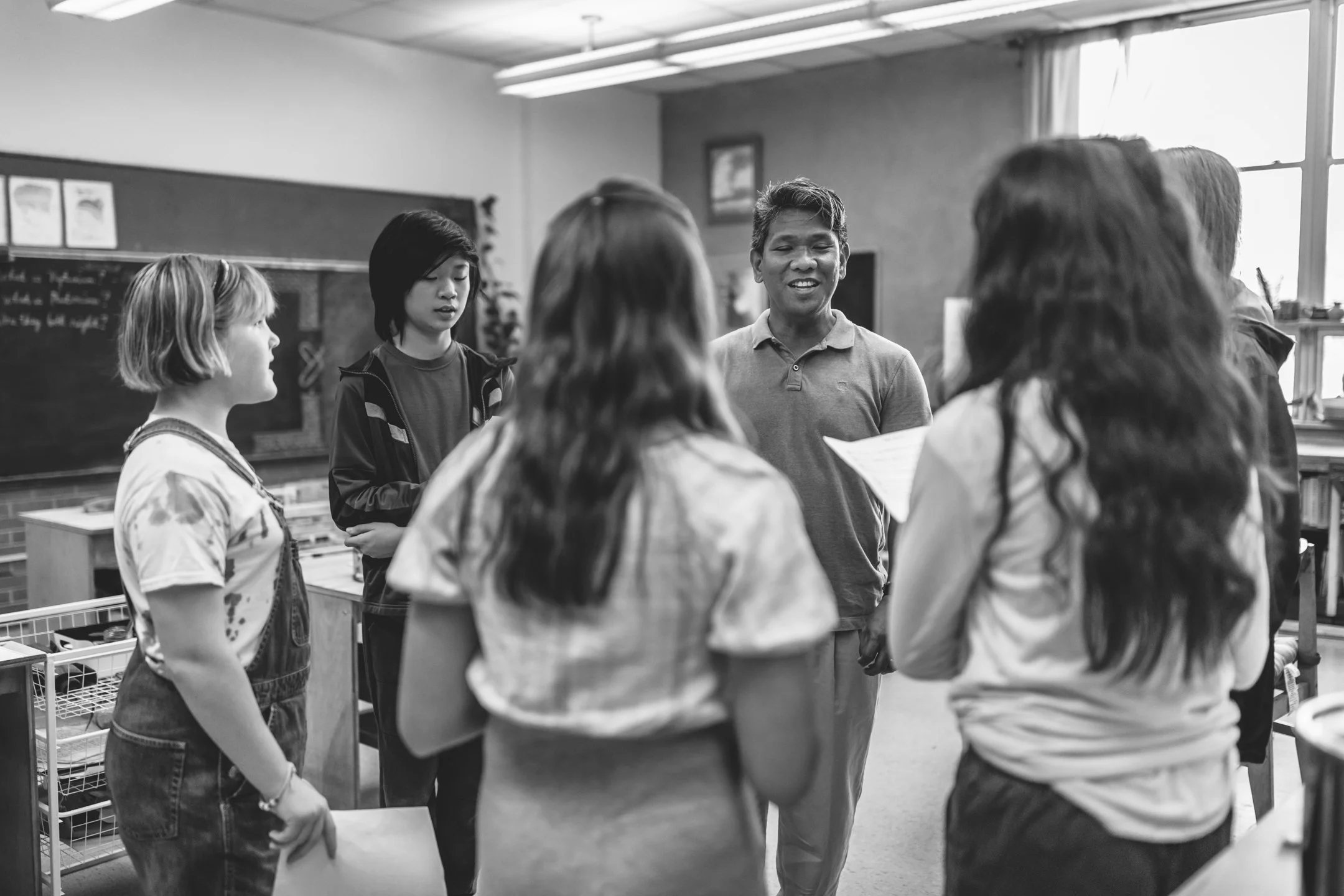 A group of four middle school students with a teacher are singing in a music class in a classroom. The students are engaged and a part of a community.