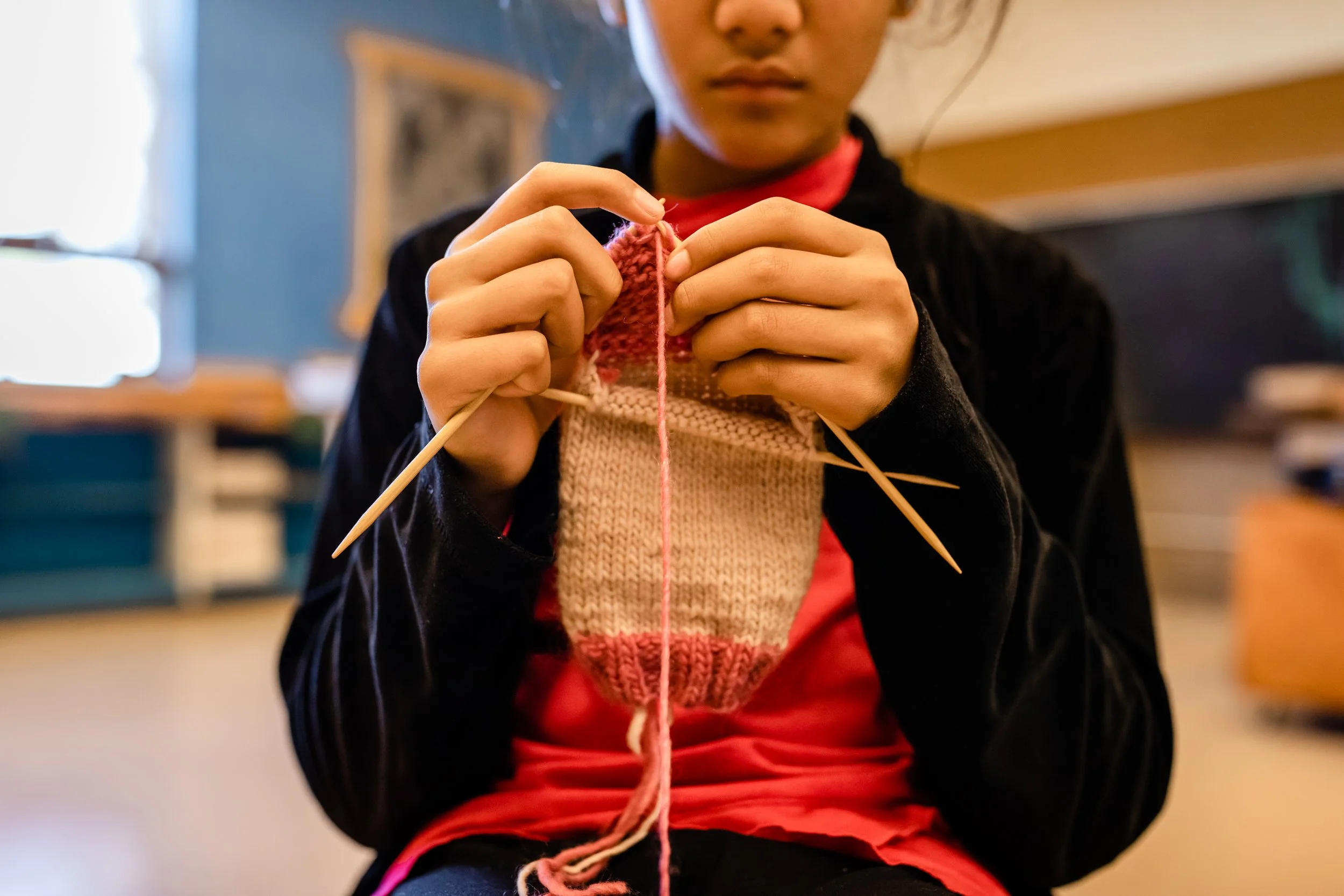 A student is sitting, knitting with three needles for a complex sock pattern. Knitting extends below the student's needles.