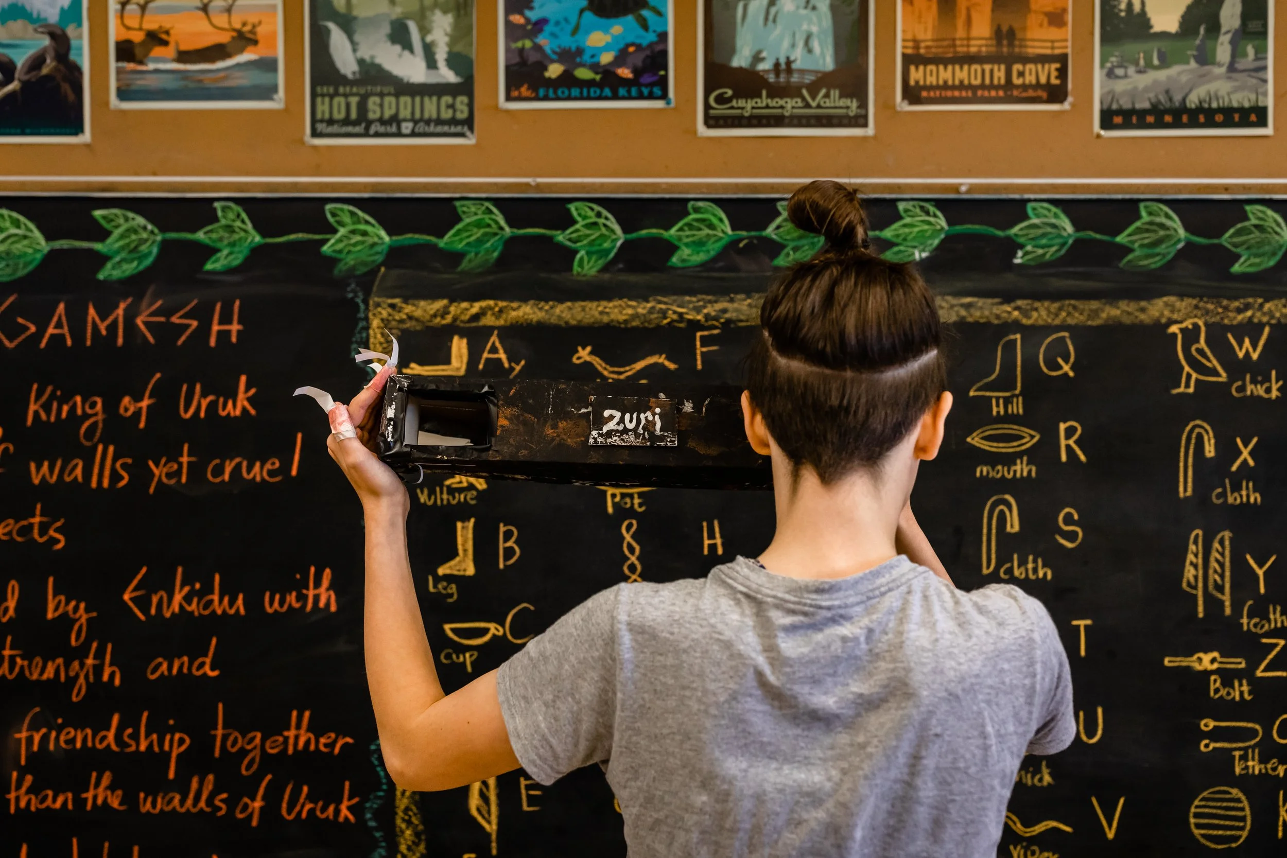 An 8th grade student stands facing a chalkboard holding a ruler in front of a handwritten world language alphabet.