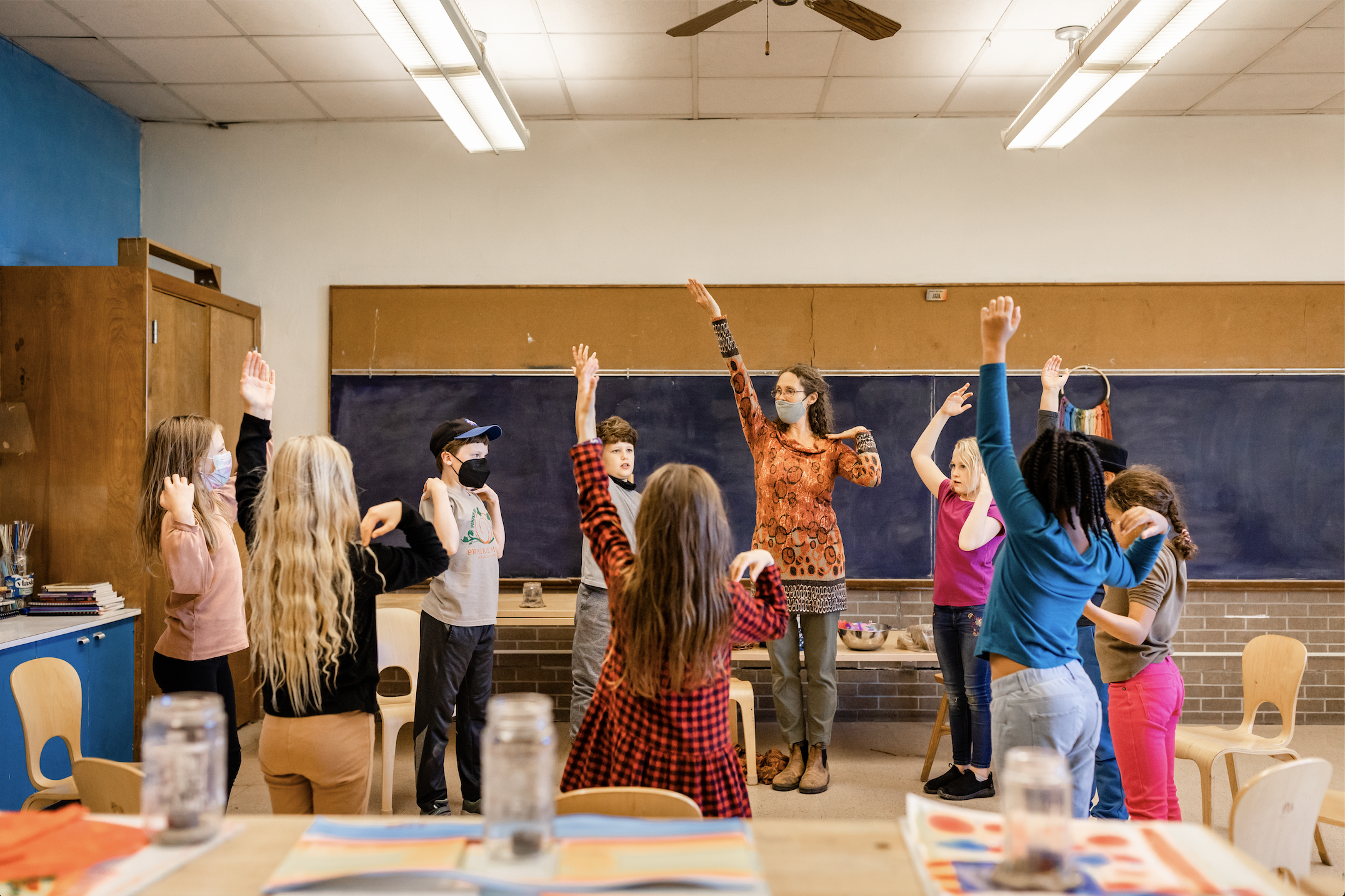 A group of students stand in a circle in a 4th grade classroom in a movement circle with their arms raised. The teacher is connected and leading the students as they follow her motions..