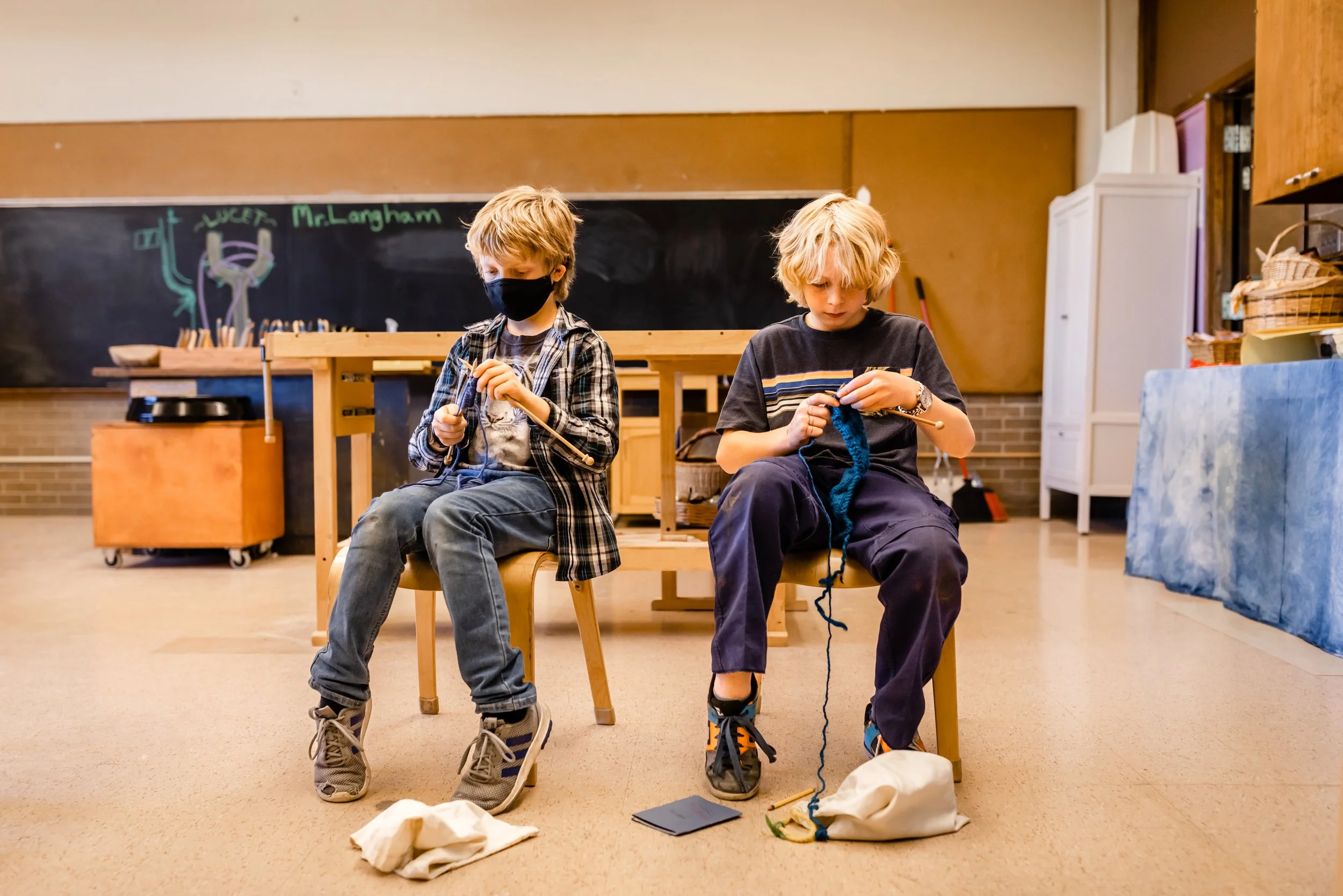 Two 5th grade students are sitting in wooden chairs in a classroom with knitting needles, knitting with blue yarn.