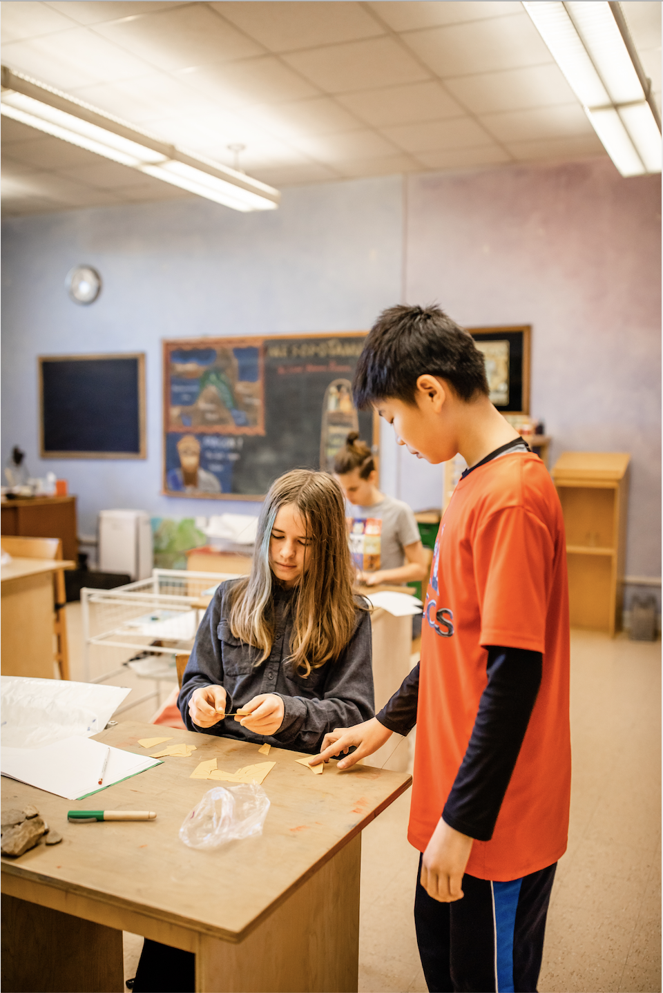 Two 6th grae students are in a classroom, one is standing, engaged with a small fragment on the table and the other is sitting, holding a small fragment. Both are engaged in hands on learning in a classroom setting in Lawrence Kansas
