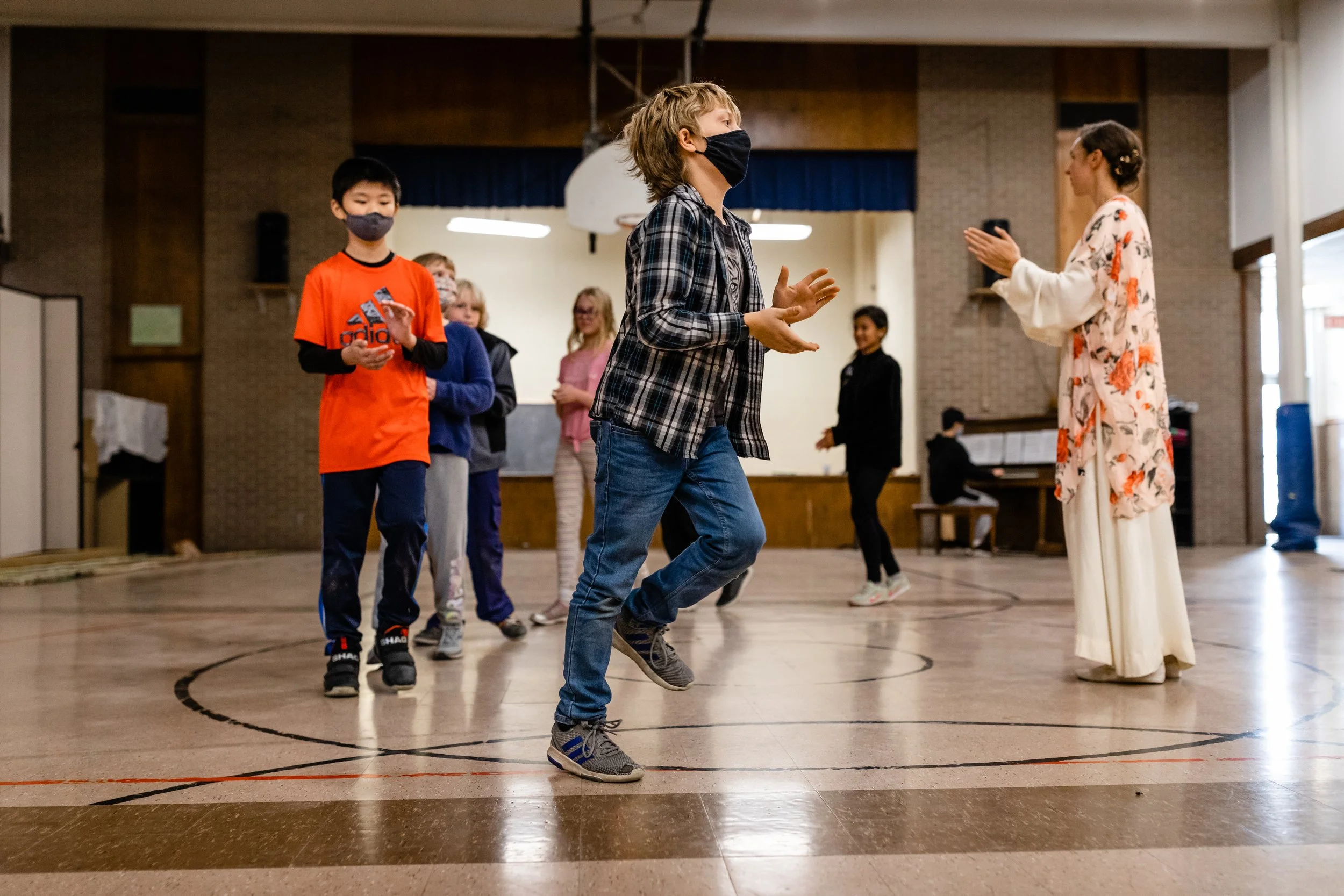 A student in a plaid shirt is skipping in forward movement while clapping as another student in an orange shirt follows. The students are in a gym and a teacher is clapping