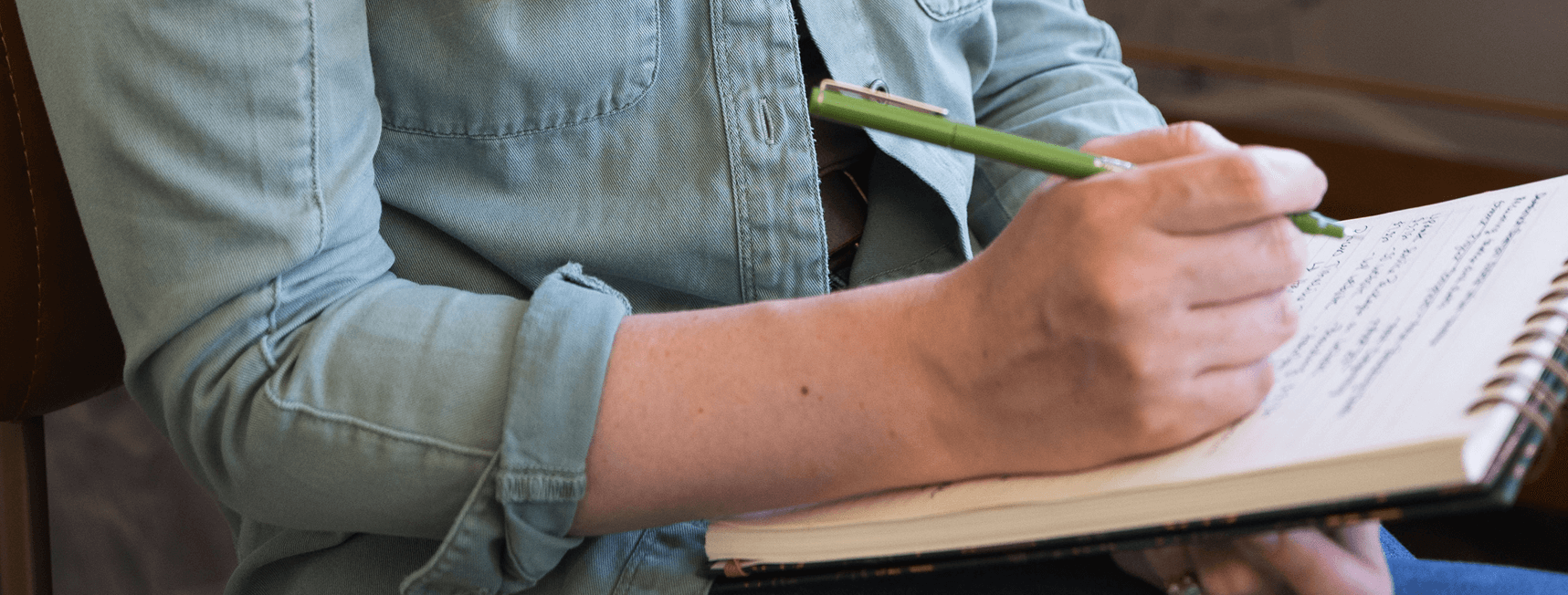 Person in a light blue denim shirt writing in a spiral notebook with a green pen.