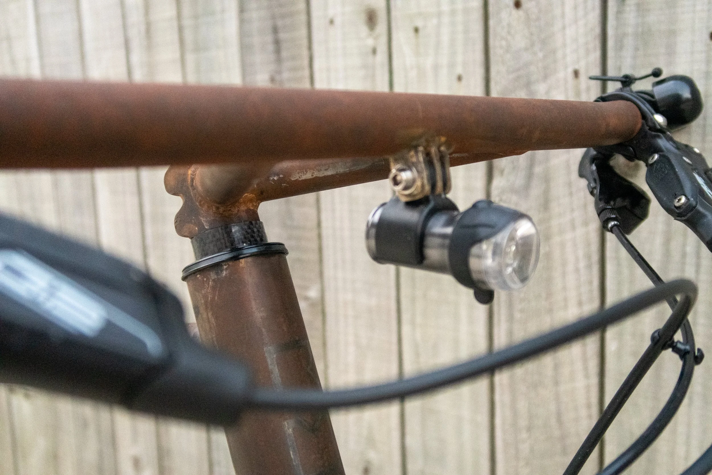 Close-up of a rusty bicycle handlebar, front light, and brake cables against a wooden fence background.