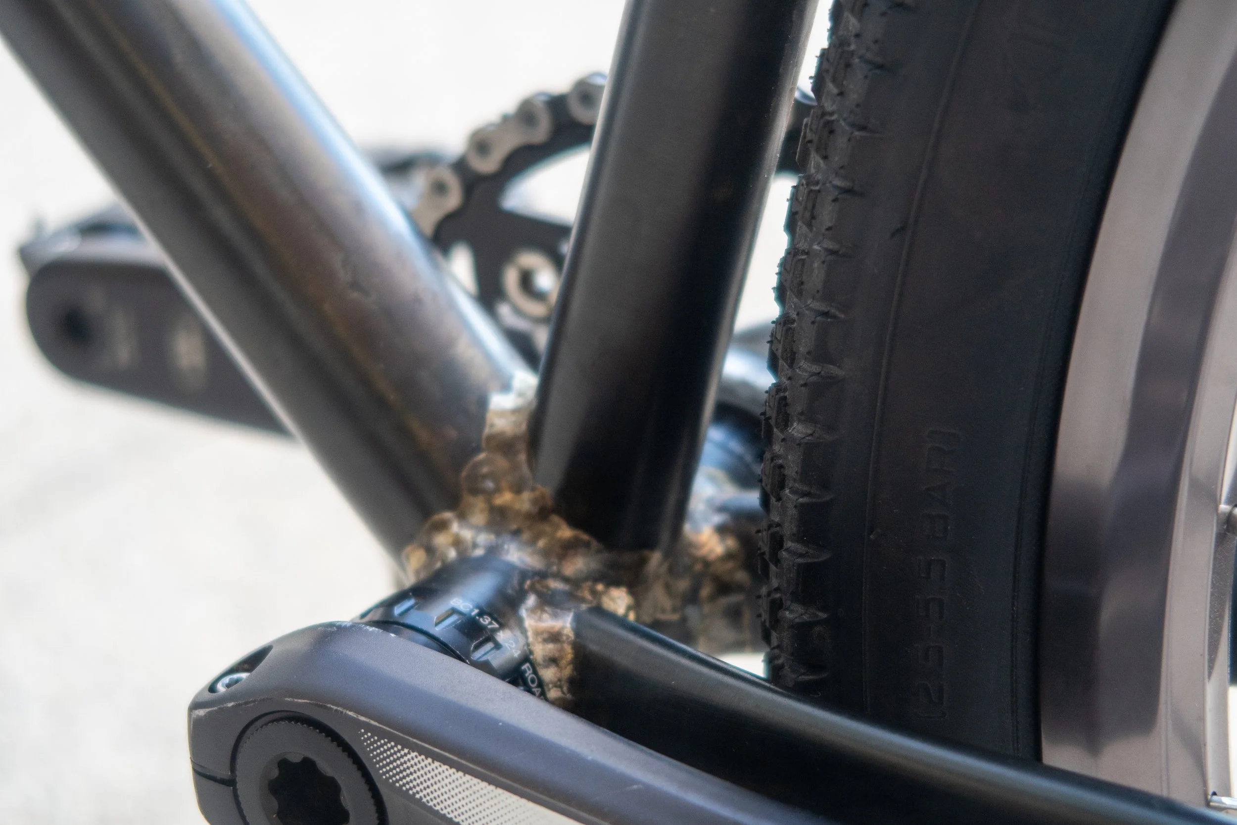 Close-up of a mountain bike's rear wheel, chain, and frame, showing some rust on the welding joint near the bottom bracket.