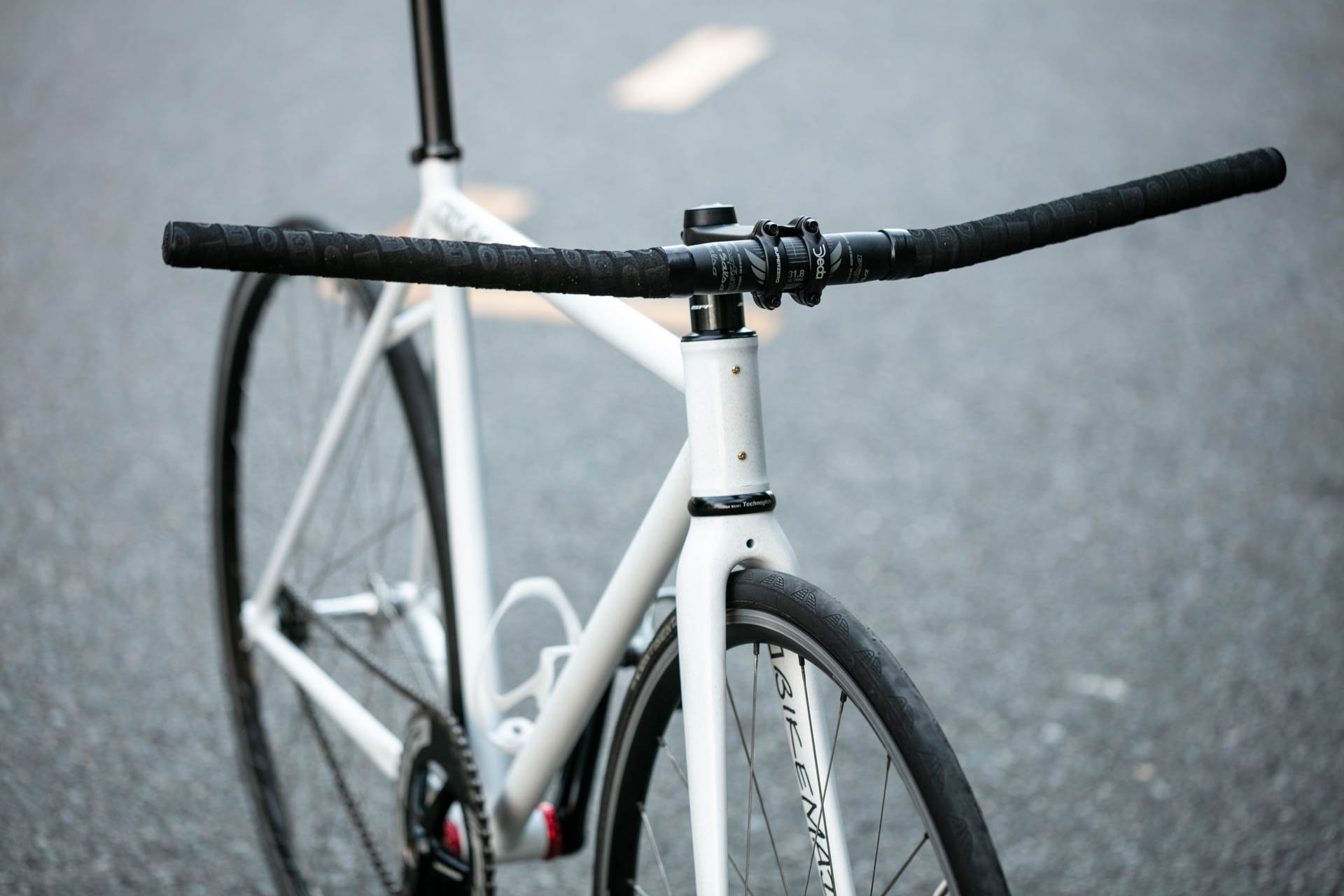 A close-up of a white road bicycle with black handlebars on a paved road.