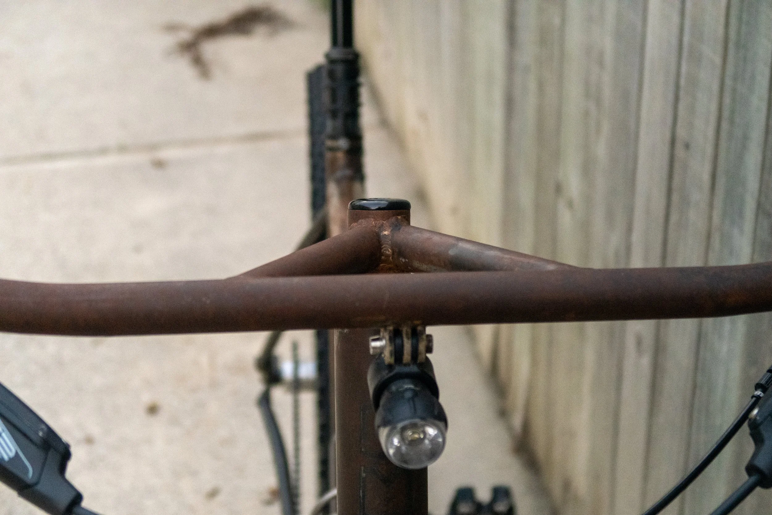 Close-up of a rusty bicycle handlebar with a front bike light attached, against a wooden fence and concrete ground.