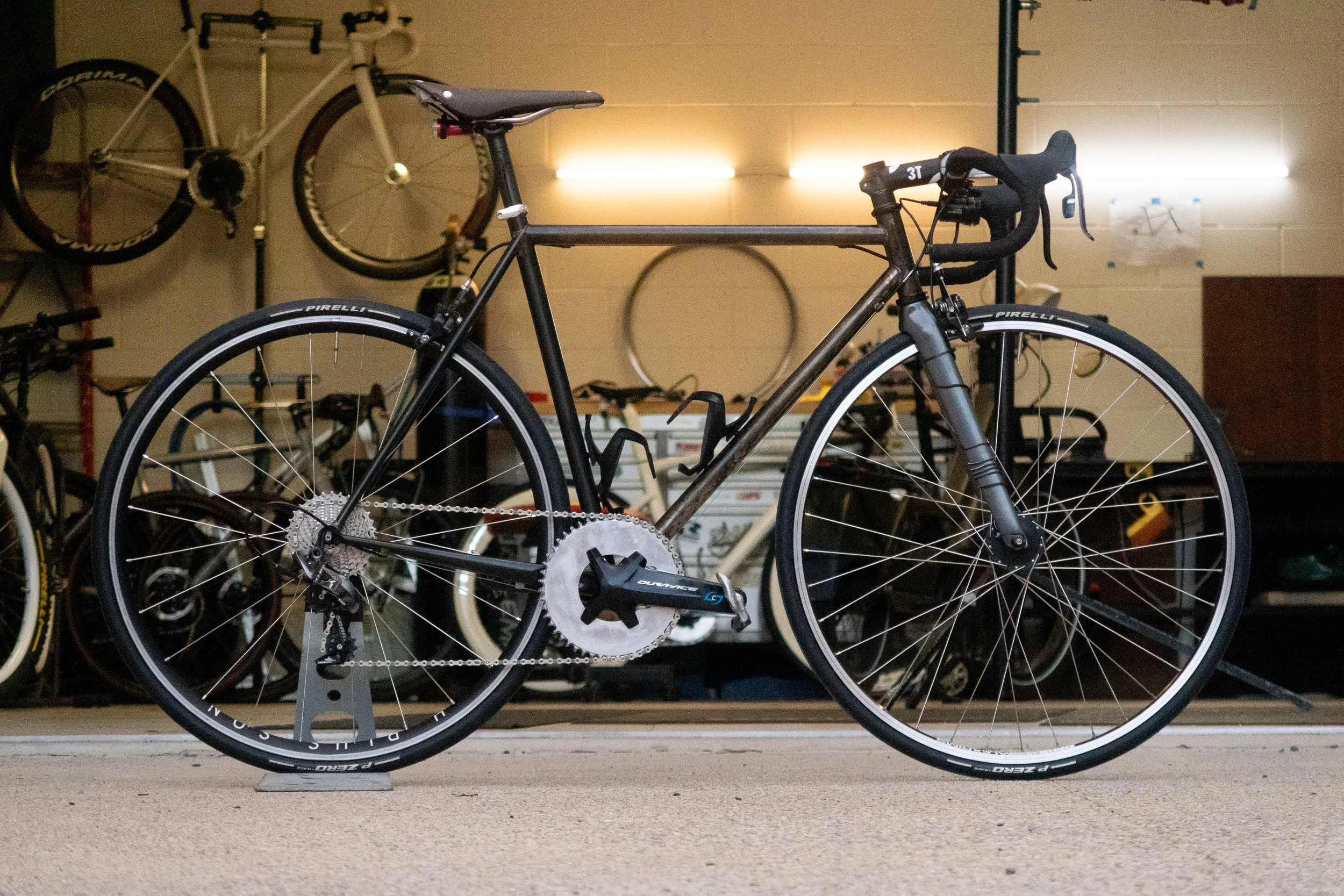 A black road bicycle with drop handlebars, disc brakes, and a Shimano Dura-Ace crankset, positioned on a bike stand indoors, with other bikes and bike accessories in the background.