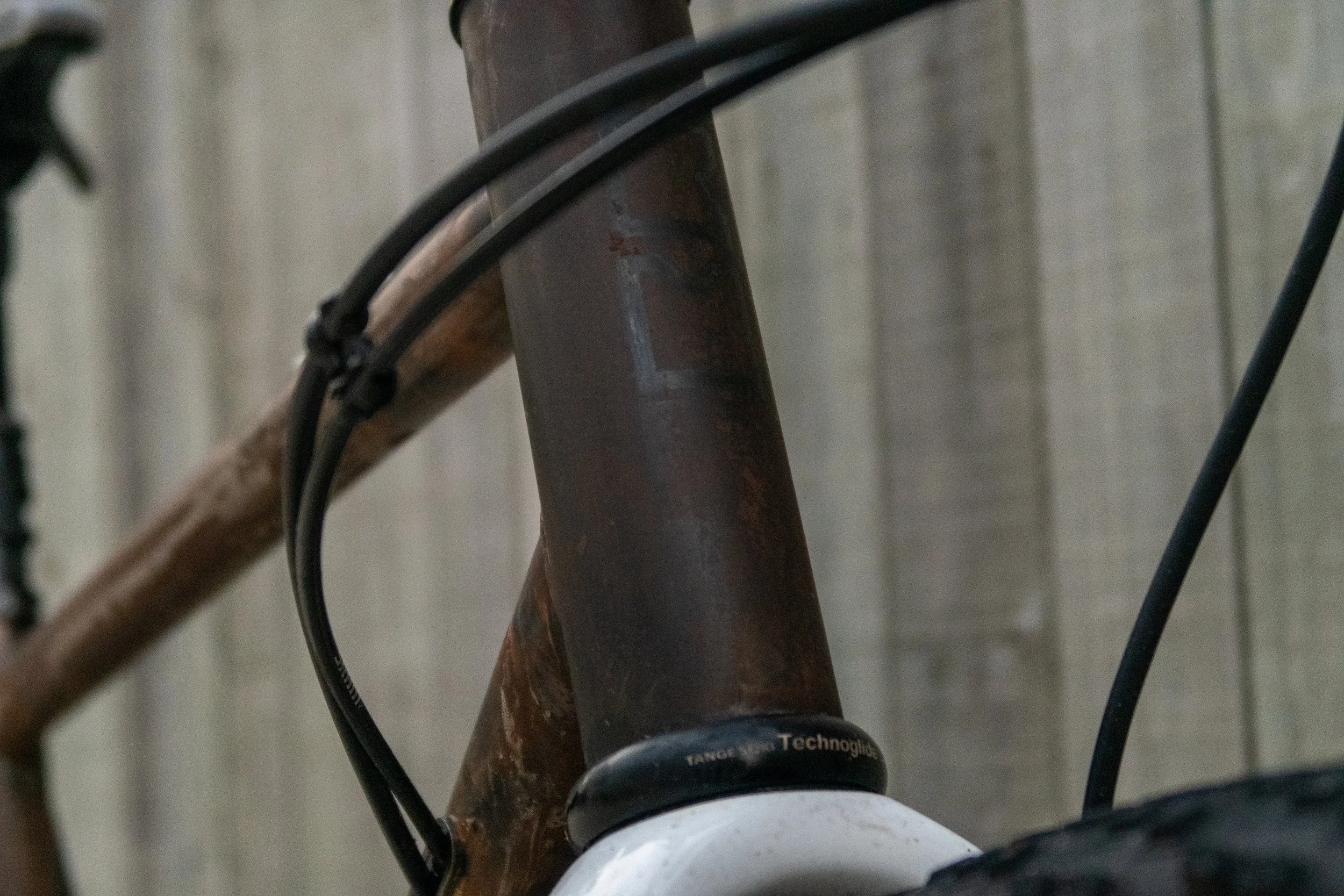Close-up of a bicycle's head tube, showing the frame with a rusted finish, cables, and part of the headset with the text 'TANGE' visible.