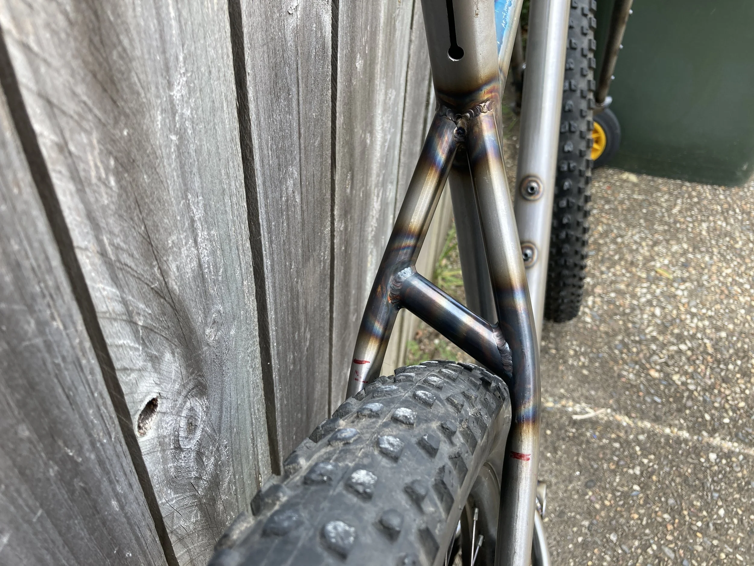 Close-up of a mountain bike's rear section showing the welded metal frame, rear tire with knobby tread, and part of the chainstay, with a wooden fence and a green trash bin in the background.