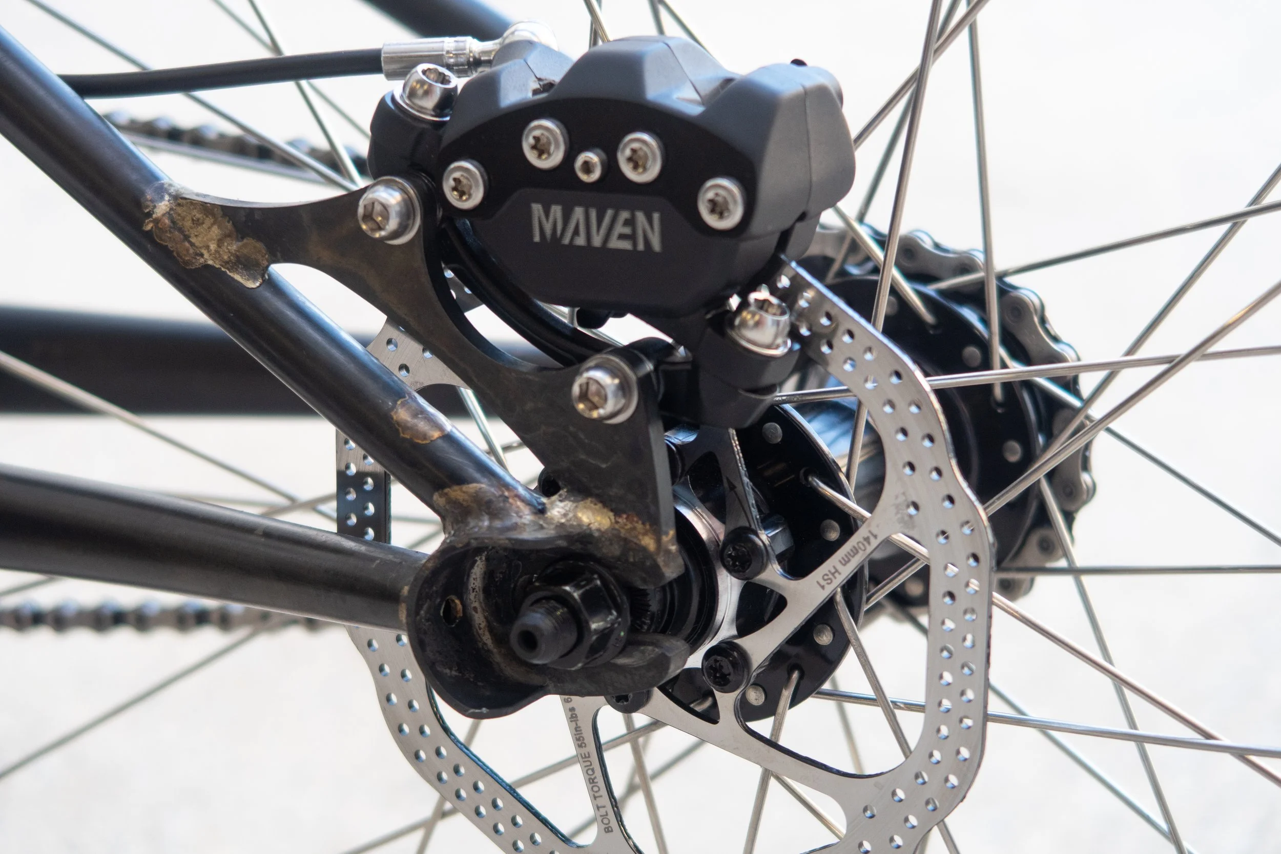 Close-up of a bicycle's rear disc brake system, showing the caliper labeled 'MAVEN,' rotor, spokes, and hub.