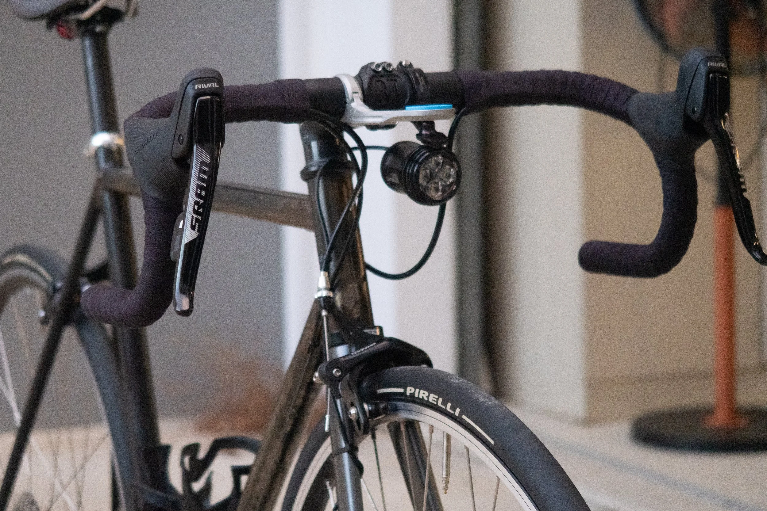 Close-up of a racing bicycle's handlebars equipped with a front light, gear shifters, and brake levers, with a blurred background.