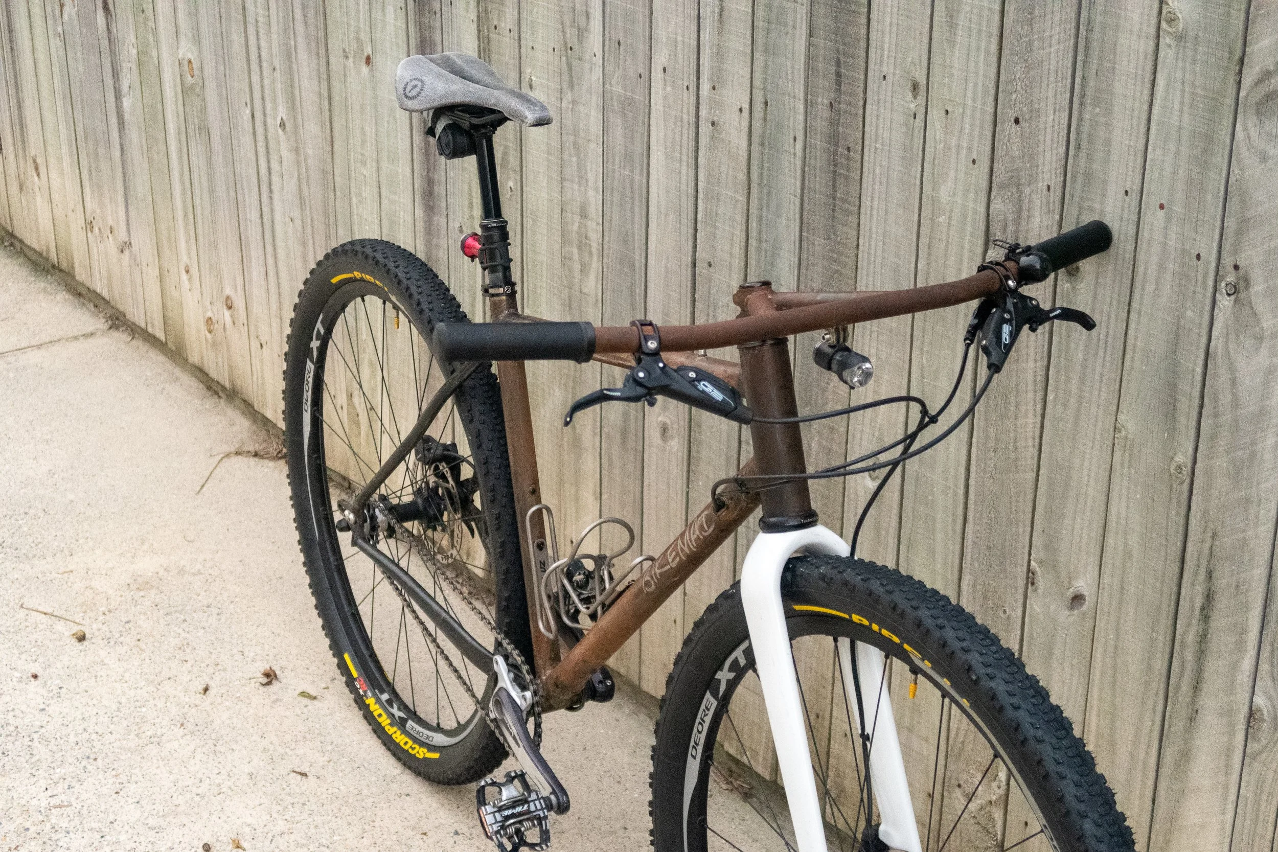 A mountain bike with a brown frame, white fork, and black tires parked against a wooden fence, with a gray saddle and black handlebars.