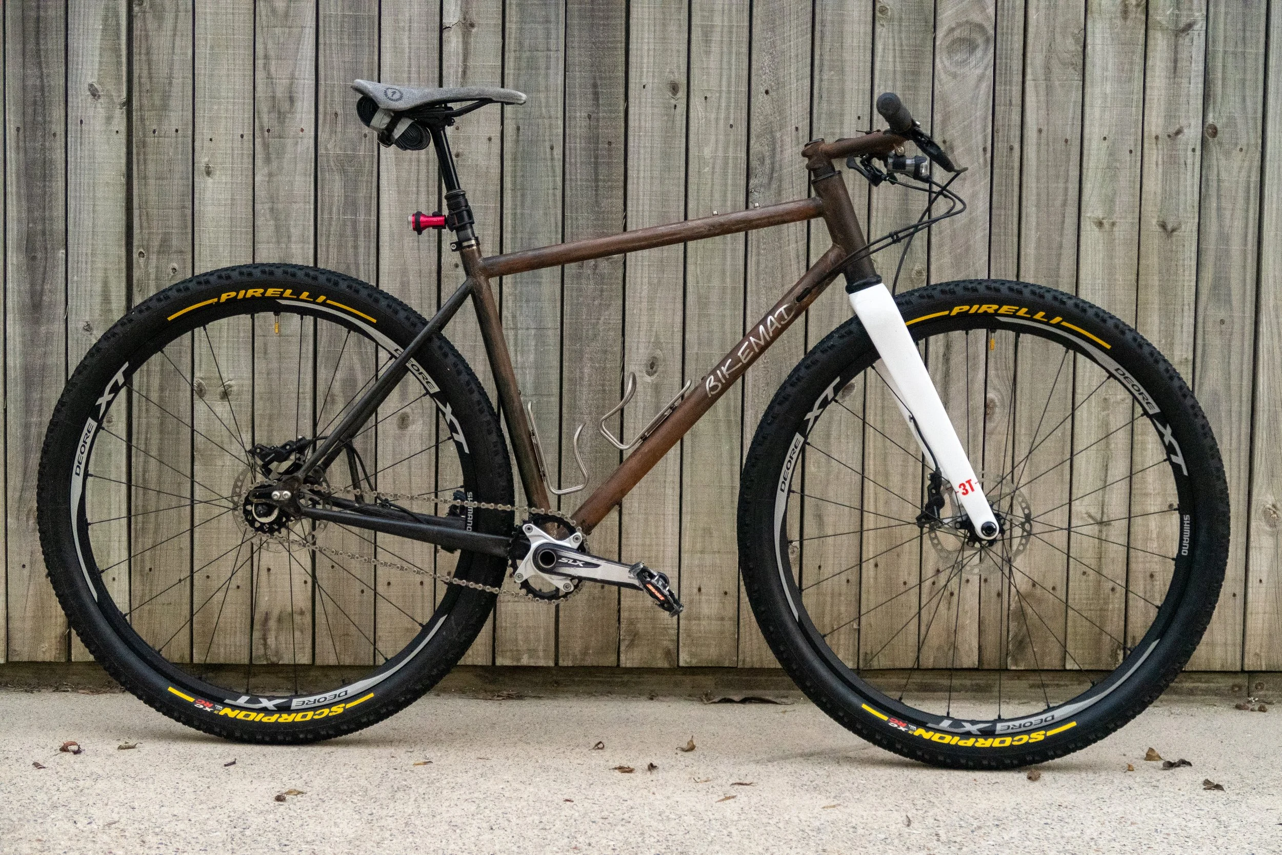 A brown mountain bike with thick tires and a white front fork, leaning against a wooden fence, with a black saddle and pedals.