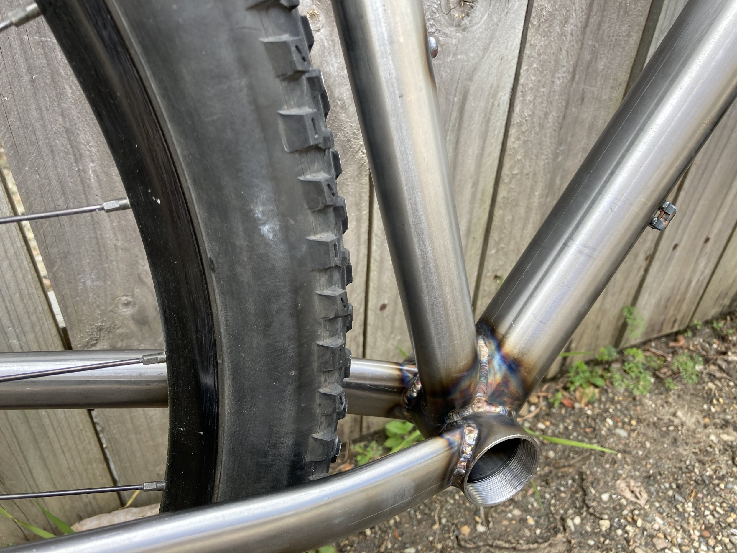 Close-up of a bicycle frame with a welded joint, stainless steel tubes, and a tire against a wooden fence background.