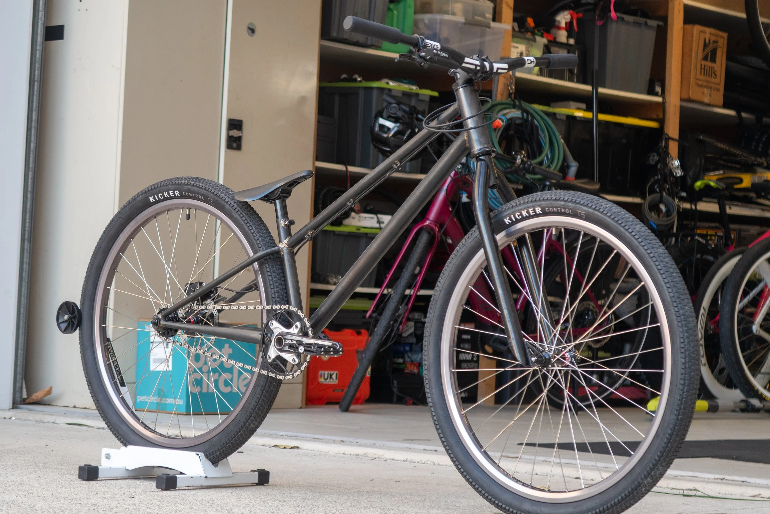 A black mountain bike with knobby tires is standing on a white bike trainer in a garage, with various tools, storage bins, and other bikes visible in the background.