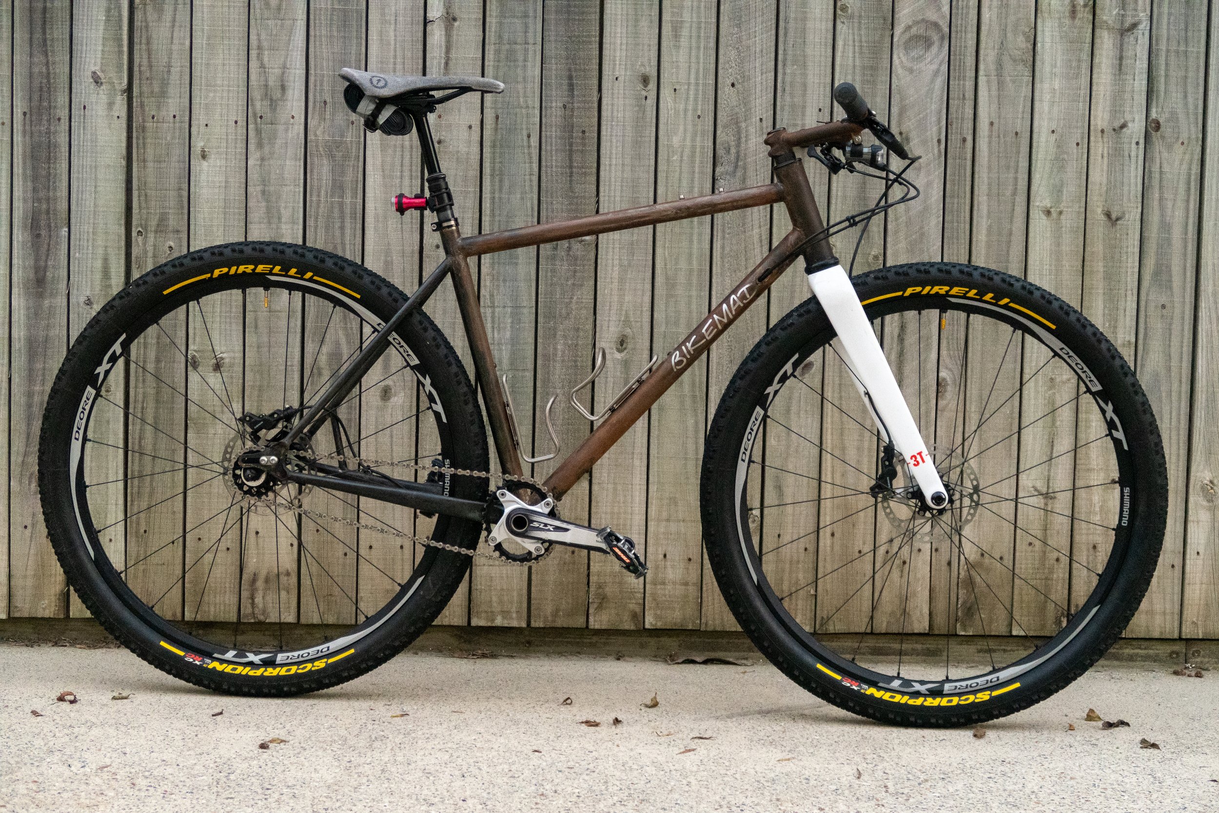 A mountain bike with a brown frame, white front fork, black tires, and yellow accents, against a wooden fence.