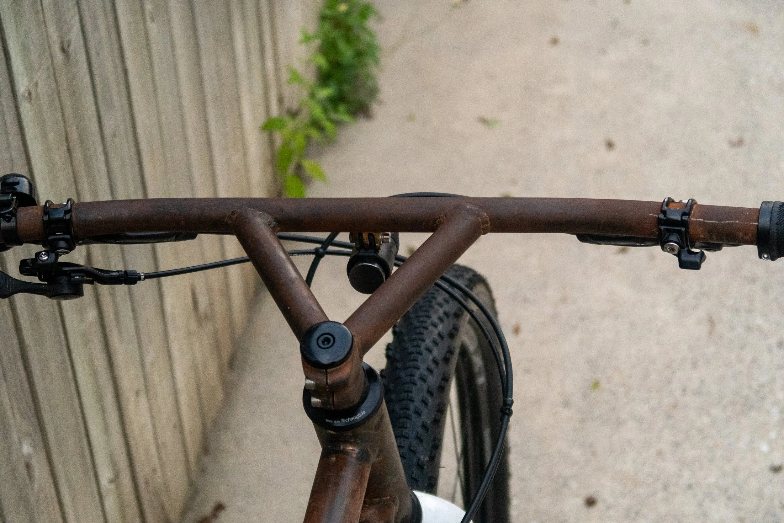 Top view of a brown mountain bike with black components, focusing on the handlebars, stem, and front tire, with a wooden fence and greenery in the background.