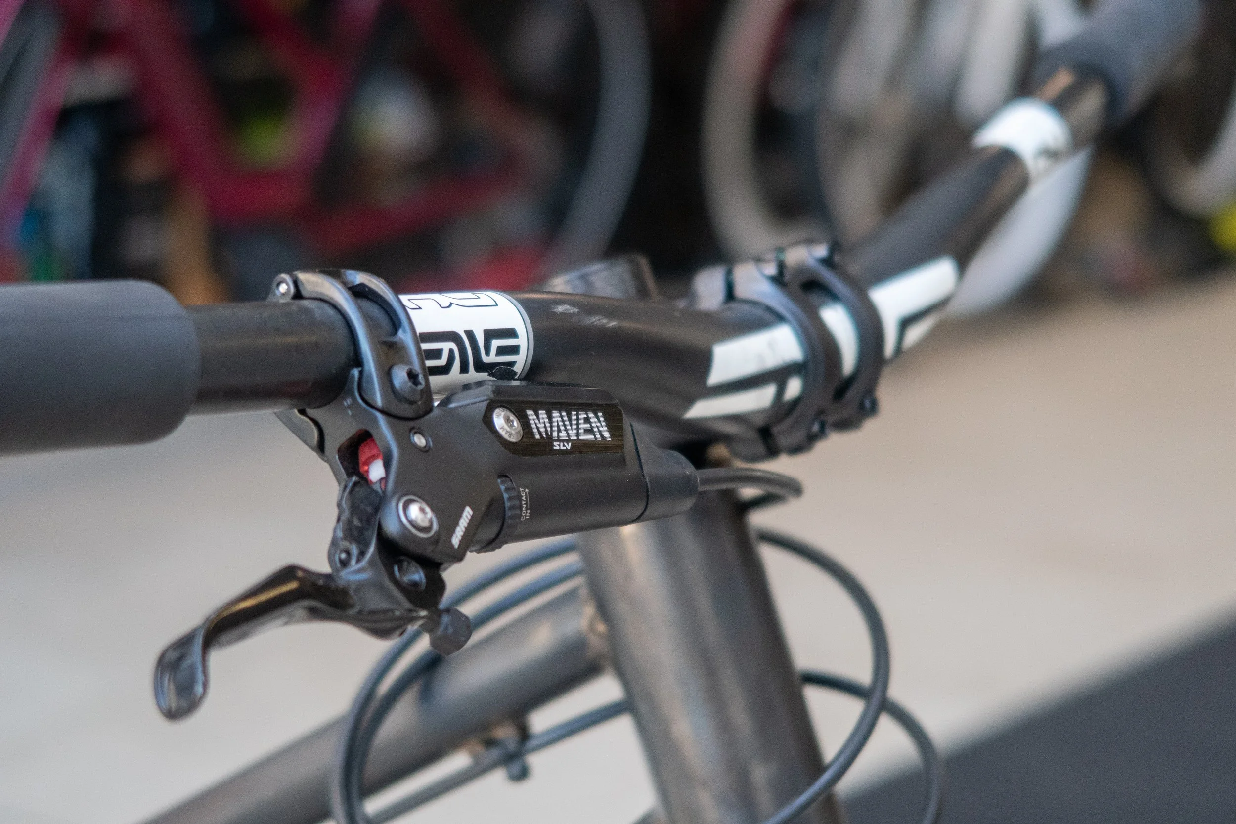 Close-up of a mountain bike handlebar with a shift sensor labeled 'MAVEN SLV' and gear shifter, with other bicycles blurred in the background.