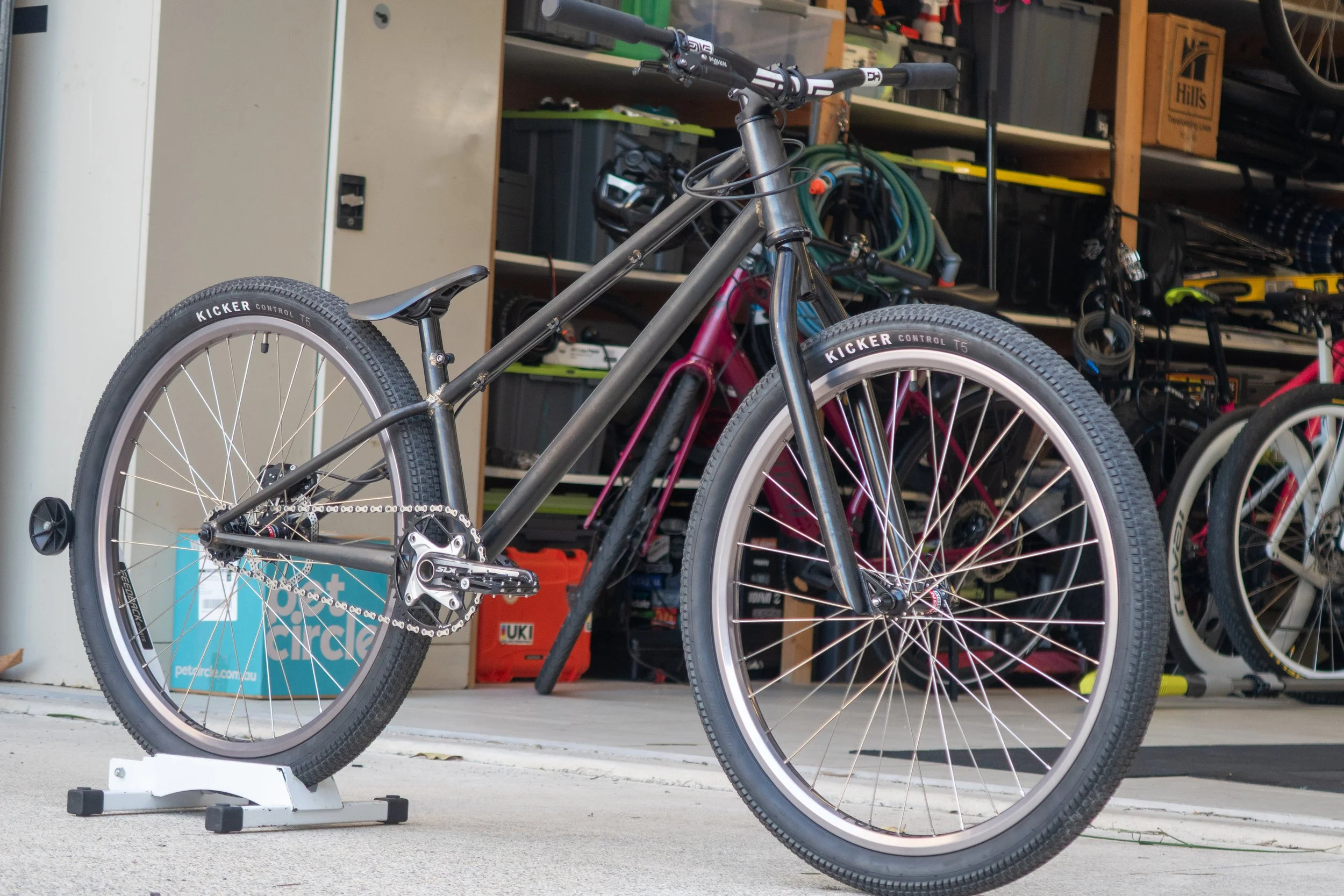 A black mountain bike with knobby tires, mounted on a white stand, inside a garage with shelves containing tools and bikes in the background.