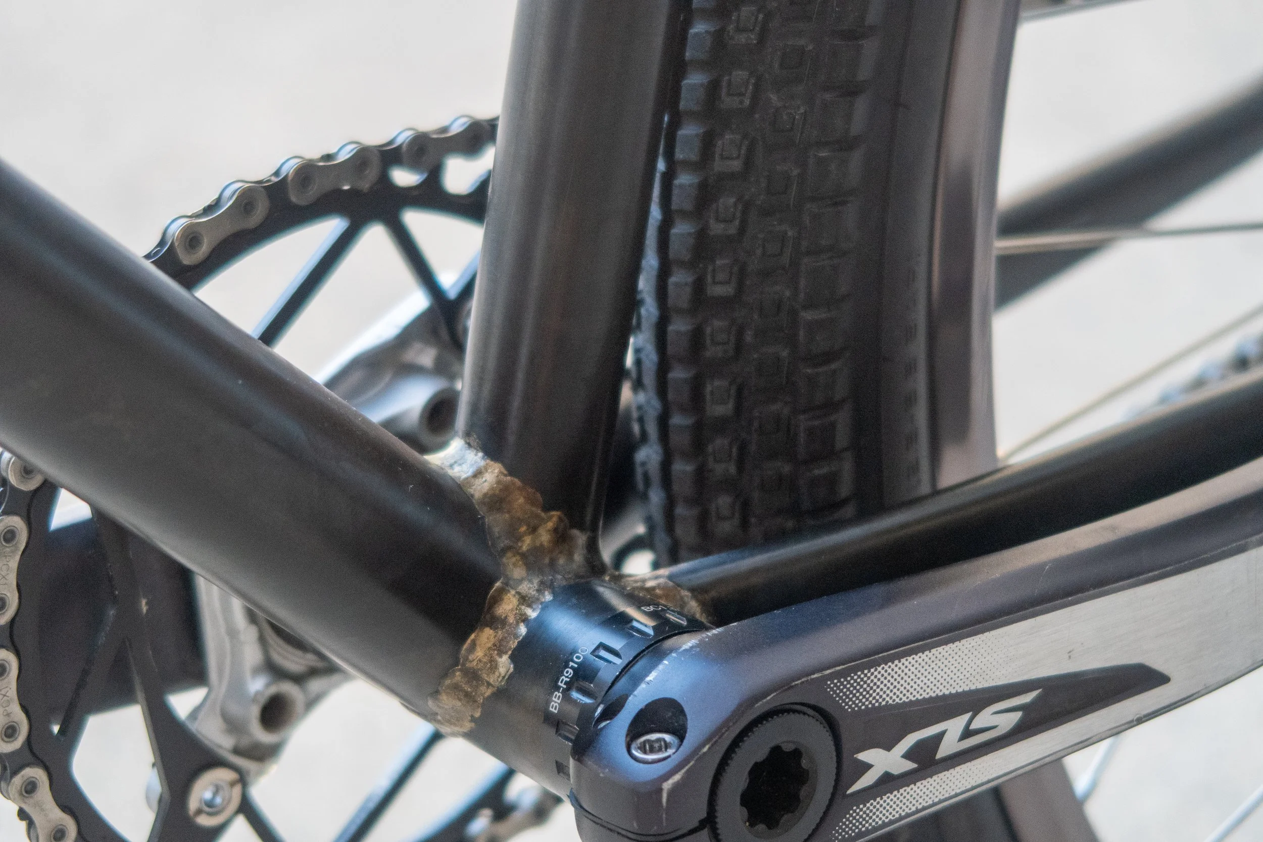 Close-up of a bicycle's bottom bracket area, showing rust on the frame and chainring, with part of the tire and pedal visible.
