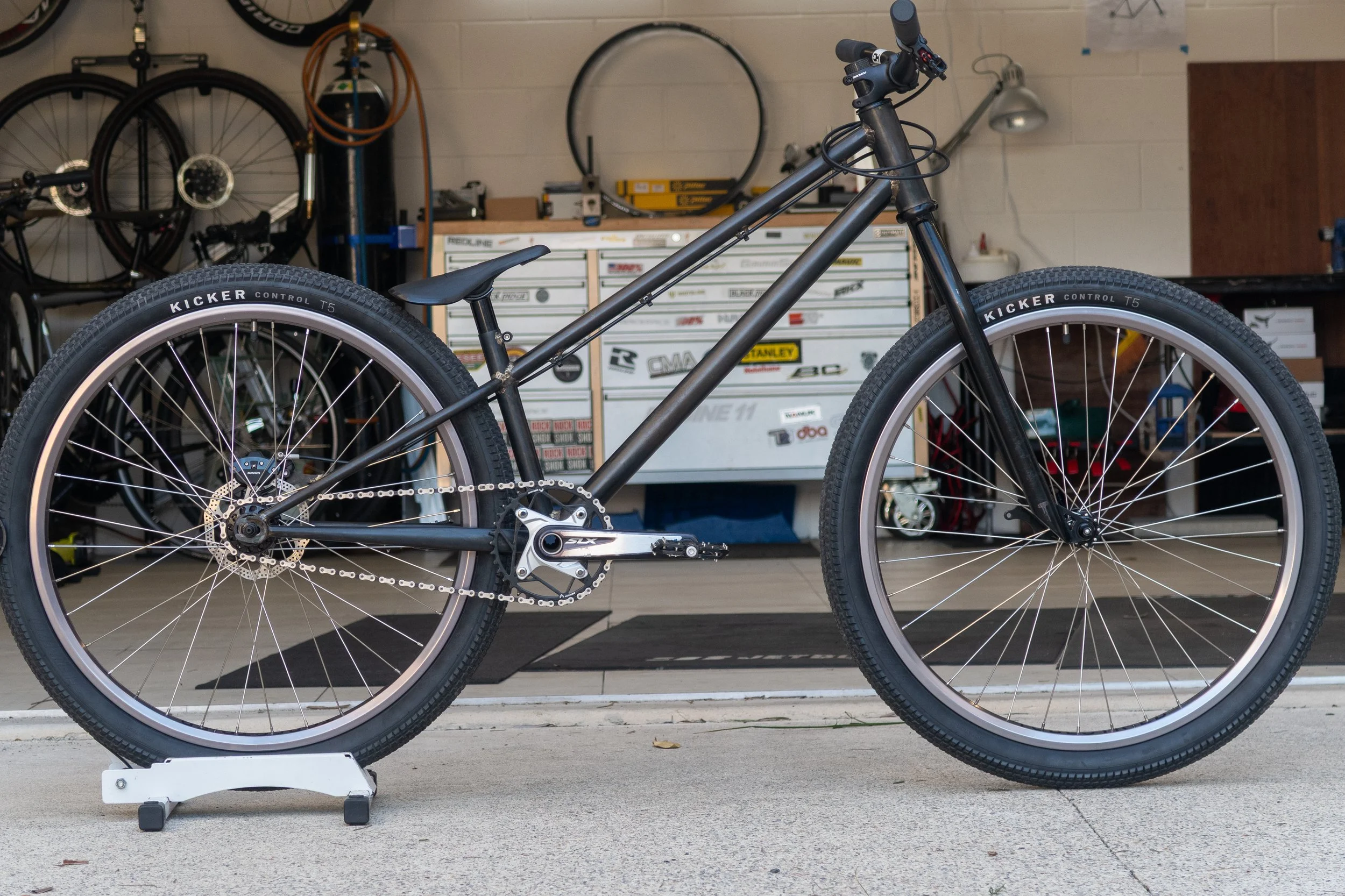 A black mountain bicycle with wide tires, resting on a bike stand in front of a garage with various tools and a tool chest in the background.